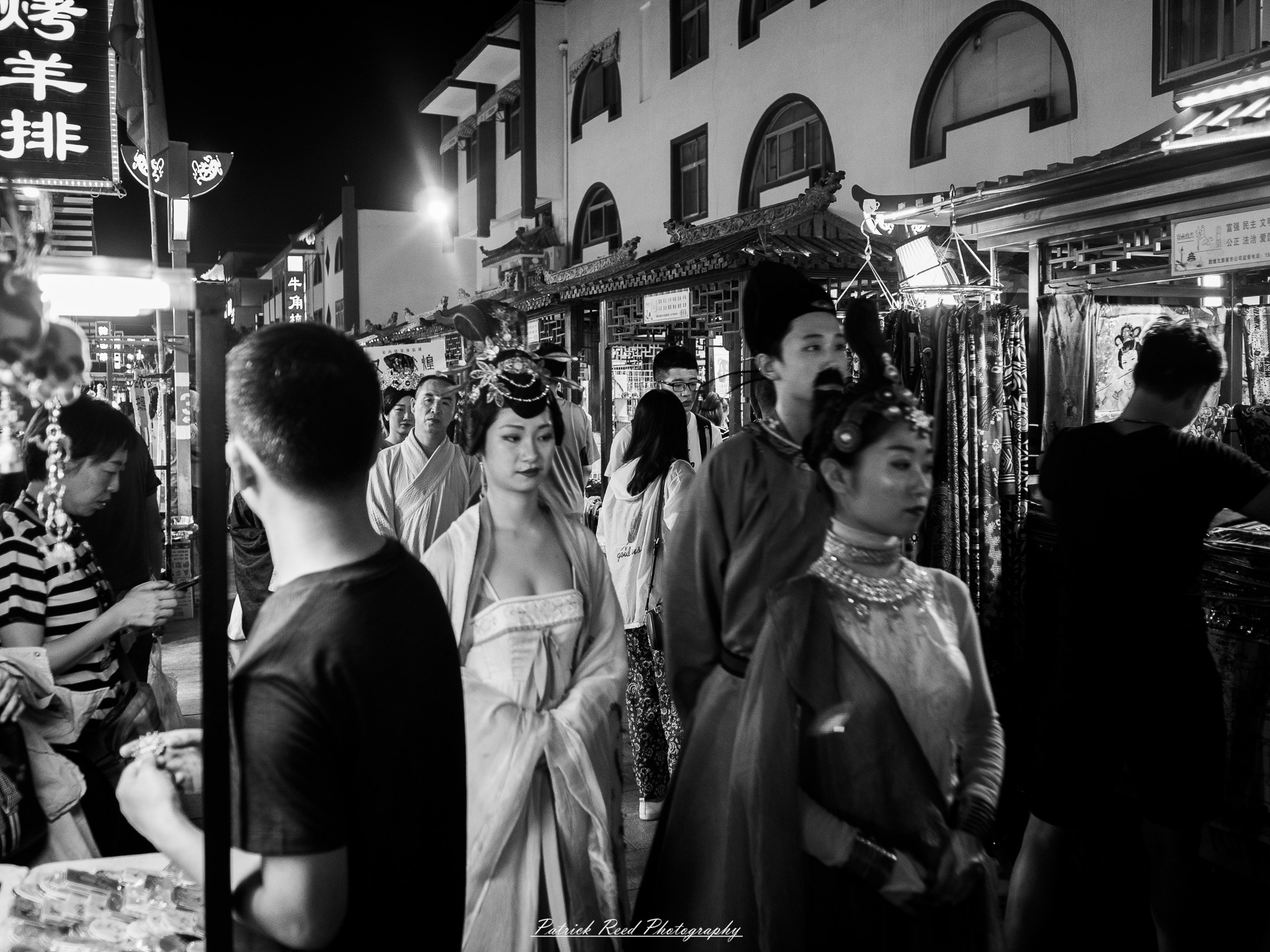 A series of noir-style street photography scenes set in Dunhuang, China, at night. The dimly lit streets and narrow alleyways reflect the soft glow of lanterns and neon signs from small shops and market stalls. Silhouettes of people walk through the misty night, some in traditional robes, adding a sense of mystery. The historic Silk Road atmosphere is highlighted by earthen architecture, intricate wooden doors, and desert dust settling on the pavement. Modern elements like motorbikes and distant headlights contrast with the ancient setting. The deep shadows and warm, flickering lights create a cinematic, moody ambiance, blending noir aesthetics with Dunhuang’s rich cultural history.