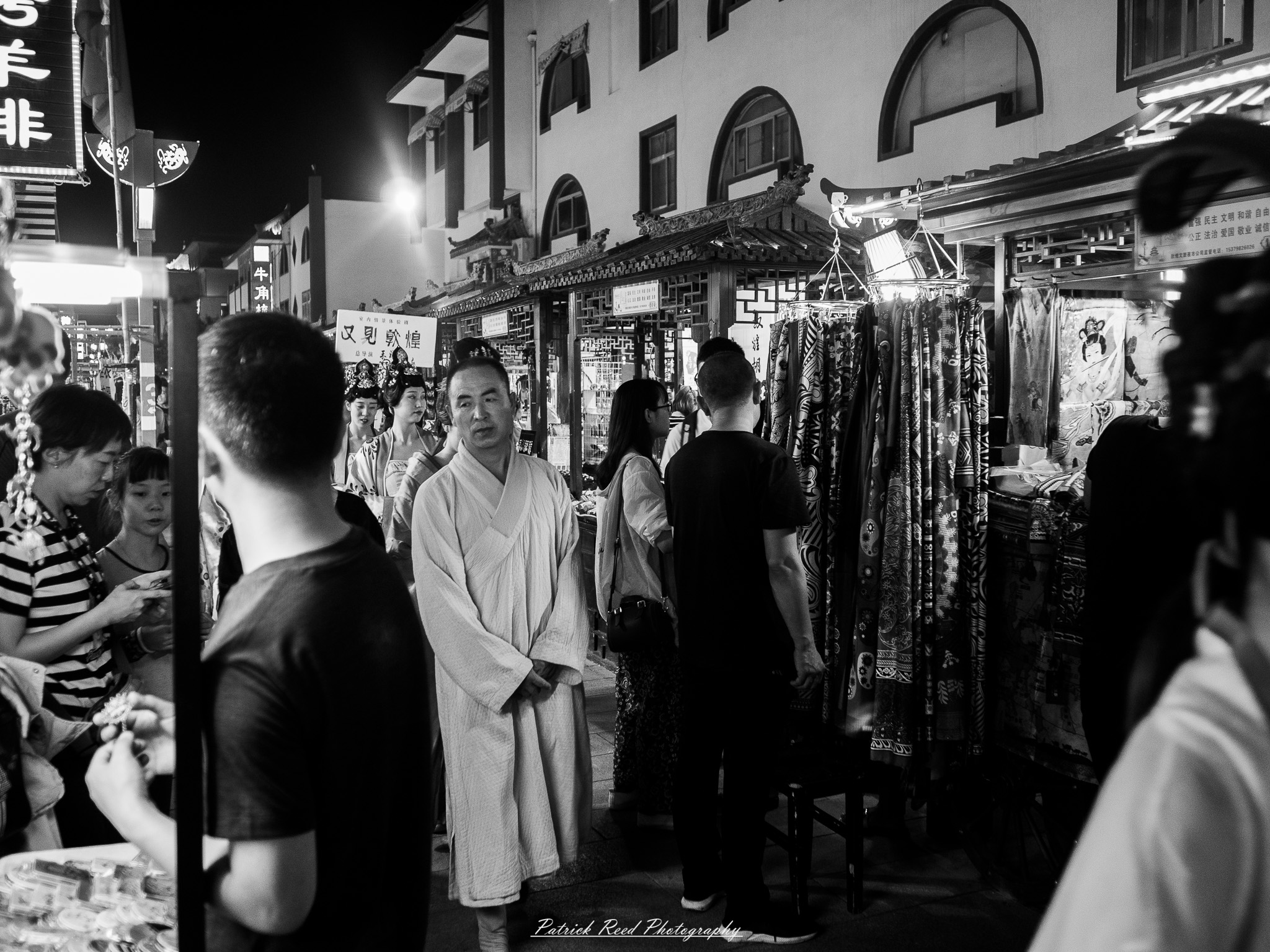 A series of noir-style street photography scenes set in Dunhuang, China, at night. The dimly lit streets and narrow alleyways reflect the soft glow of lanterns and neon signs from small shops and market stalls. Silhouettes of people walk through the misty night, some in traditional robes, adding a sense of mystery. The historic Silk Road atmosphere is highlighted by earthen architecture, intricate wooden doors, and desert dust settling on the pavement. Modern elements like motorbikes and distant headlights contrast with the ancient setting. The deep shadows and warm, flickering lights create a cinematic, moody ambiance, blending noir aesthetics with Dunhuang’s rich cultural history.