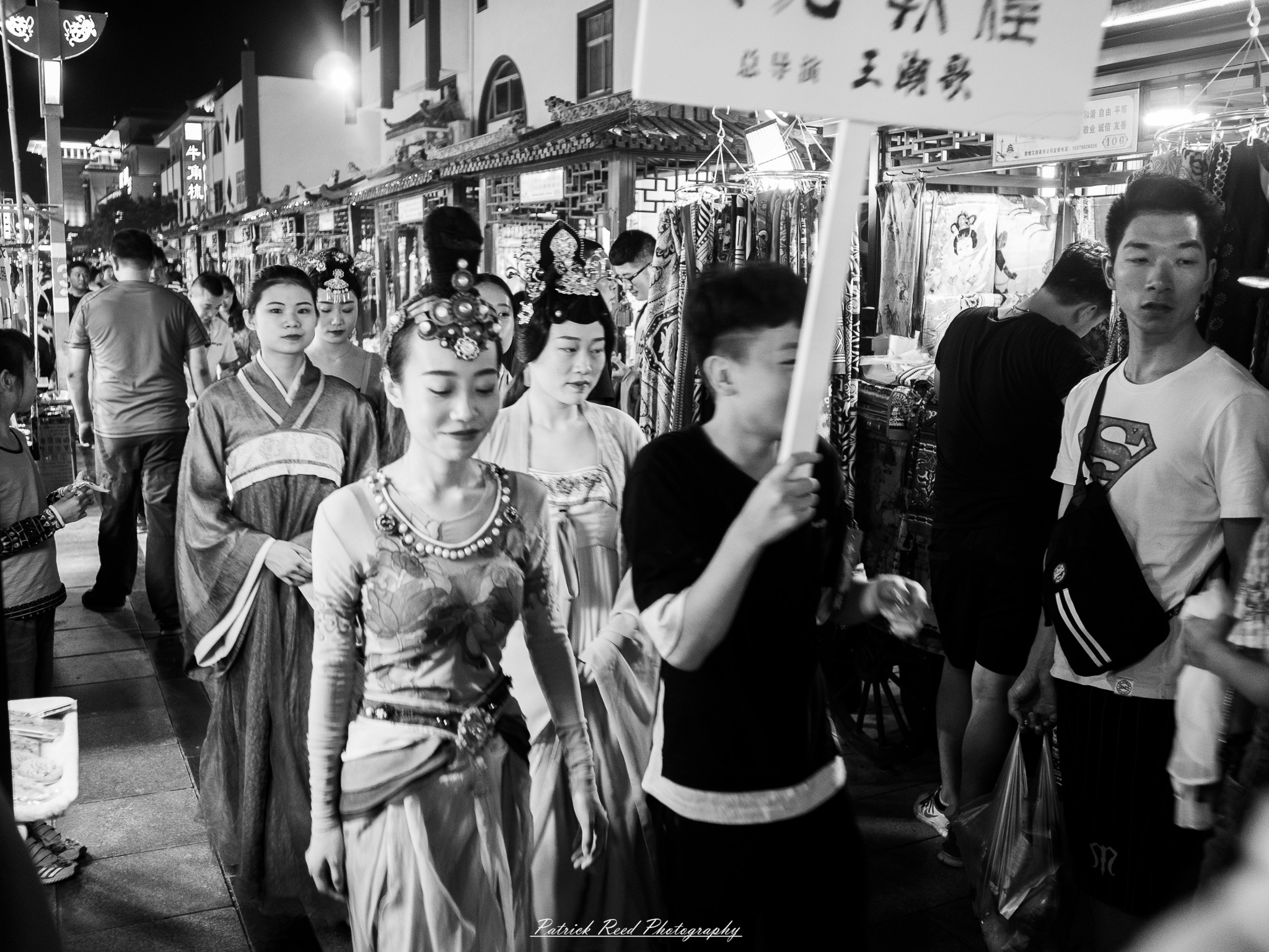 Dunhuang 4 A series of noir-style street photography scenes set in Dunhuang, China, at night. The dimly lit streets and narrow alleyways reflect the soft glow of lanterns and neon signs from small shops and market stalls. Silhouettes of people walk through the misty night, some in traditional robes, adding a sense of mystery. The historic Silk Road atmosphere is highlighted by earthen architecture, intricate wooden doors, and desert dust settling on the pavement. Modern elements like motorbikes and distant headlights contrast with the ancient setting. The deep shadows and warm, flickering lights create a cinematic, moody ambiance, blending noir aesthetics with Dunhuang’s rich cultural history.