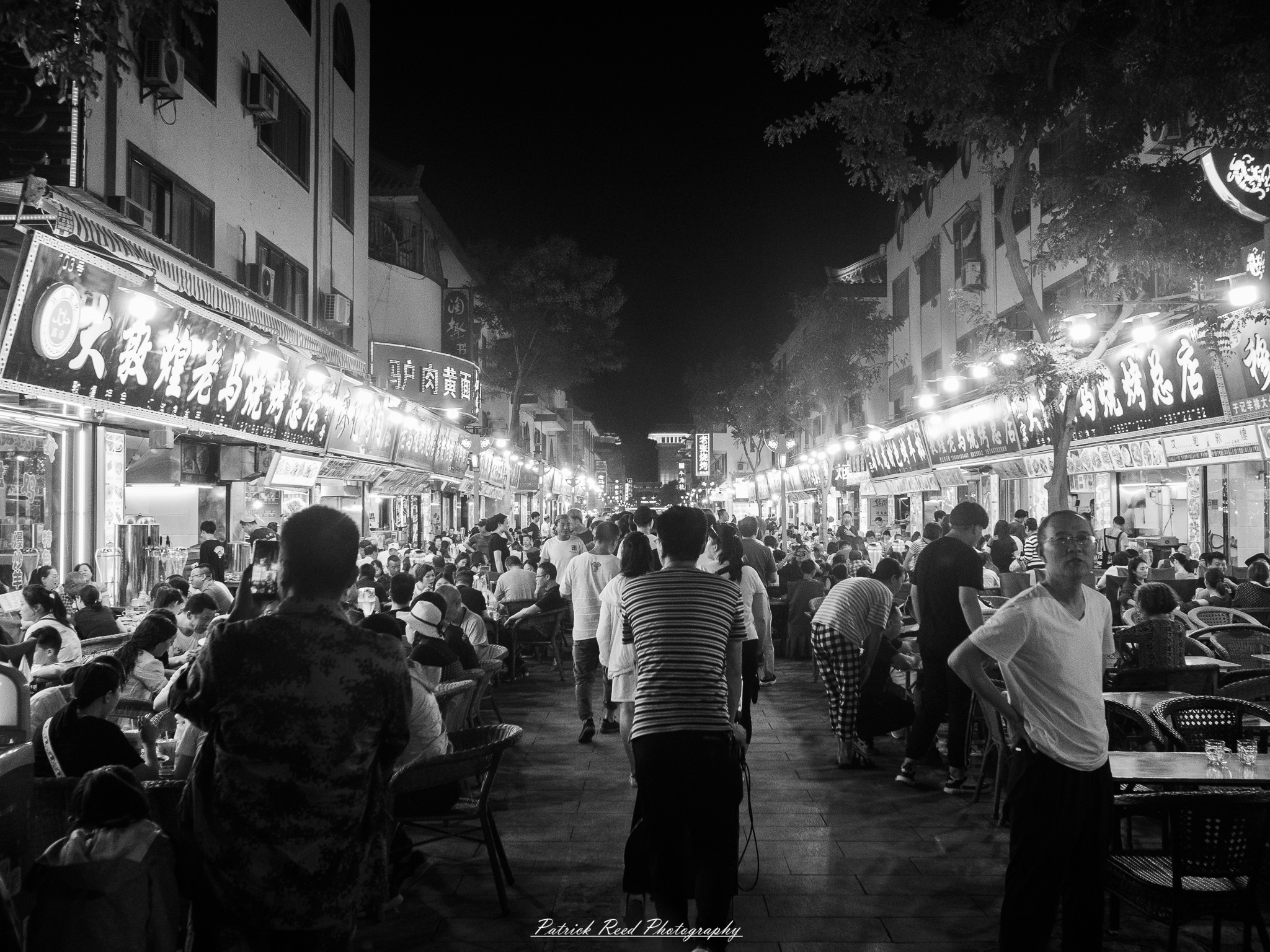 A series of noir-style street photography scenes set in Dunhuang, China, at night. The dimly lit streets and narrow alleyways reflect the soft glow of lanterns and neon signs from small shops and market stalls. Silhouettes of people walk through the misty night, some in traditional robes, adding a sense of mystery. The historic Silk Road atmosphere is highlighted by earthen architecture, intricate wooden doors, and desert dust settling on the pavement. Modern elements like motorbikes and distant headlights contrast with the ancient setting. The deep shadows and warm, flickering lights create a cinematic, moody ambiance, blending noir aesthetics with Dunhuang’s rich cultural history.