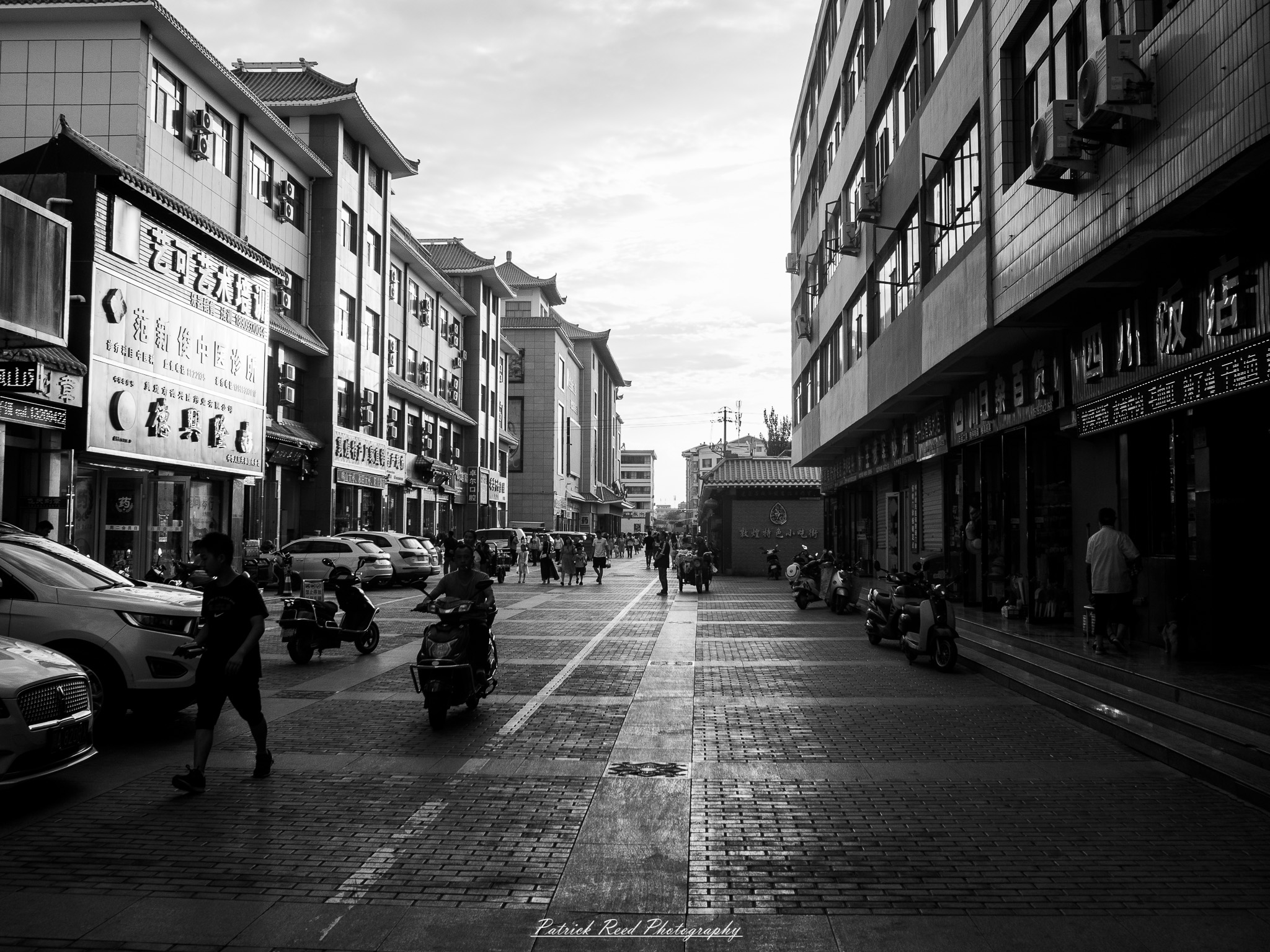 A series of noir-style street photography scenes set in Dunhuang, China, at night. The dimly lit streets and narrow alleyways reflect the soft glow of lanterns and neon signs from small shops and market stalls. Silhouettes of people walk through the misty night, some in traditional robes, adding a sense of mystery. The historic Silk Road atmosphere is highlighted by earthen architecture, intricate wooden doors, and desert dust settling on the pavement. Modern elements like motorbikes and distant headlights contrast with the ancient setting. The deep shadows and warm, flickering lights create a cinematic, moody ambiance, blending noir aesthetics with Dunhuang’s rich cultural history.