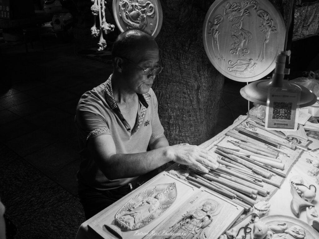 A series of noir-style street photography scenes set in Dunhuang, China, at night. The dimly lit streets and narrow alleyways reflect the soft glow of lanterns and neon signs from small shops and market stalls. Silhouettes of people walk through the misty night, some in traditional robes, adding a sense of mystery. The historic Silk Road atmosphere is highlighted by earthen architecture, intricate wooden doors, and desert dust settling on the pavement. Modern elements like motorbikes and distant headlights contrast with the ancient setting. The deep shadows and warm, flickering lights create a cinematic, moody ambiance, blending noir aesthetics with Dunhuang’s rich cultural history.