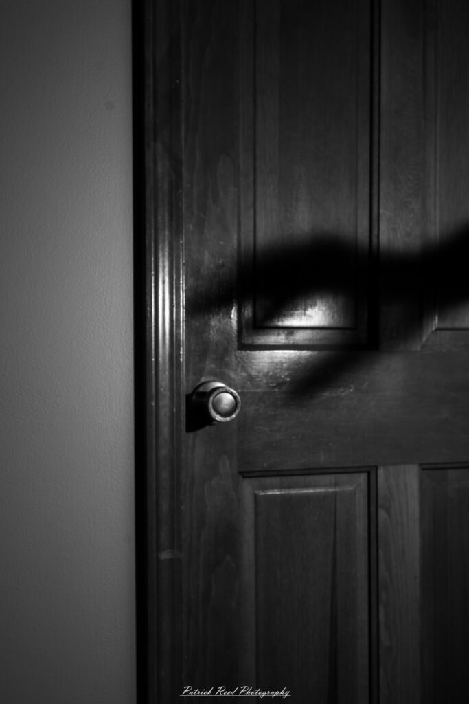 A shadowy, indistinct hand grips a door handle, its eerie silhouette contrasting against the dimly lit background. The fingers appear elongated and unnatural, creating a sense of mystery and unease.