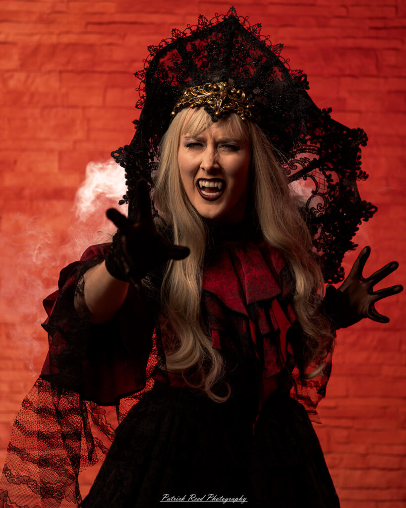 Alt text: Portrait of a woman in vampire cosplay named Lady Thorn, photographed on June 28, 2025. She is posed against a vivid red background, styled with gothic elements and dramatic expression.