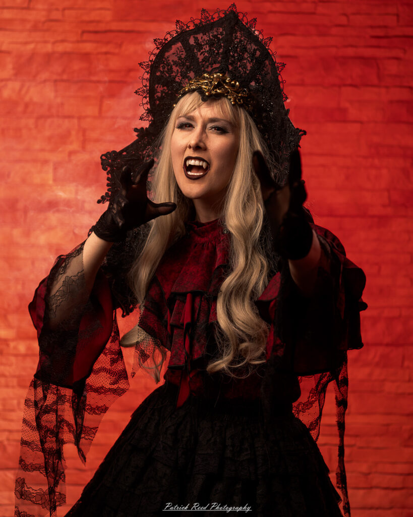 Alt text: Portrait of a woman in vampire cosplay named Lady Thorn, photographed on June 28, 2025. She is posed against a vivid red background, styled with gothic elements and dramatic expression.