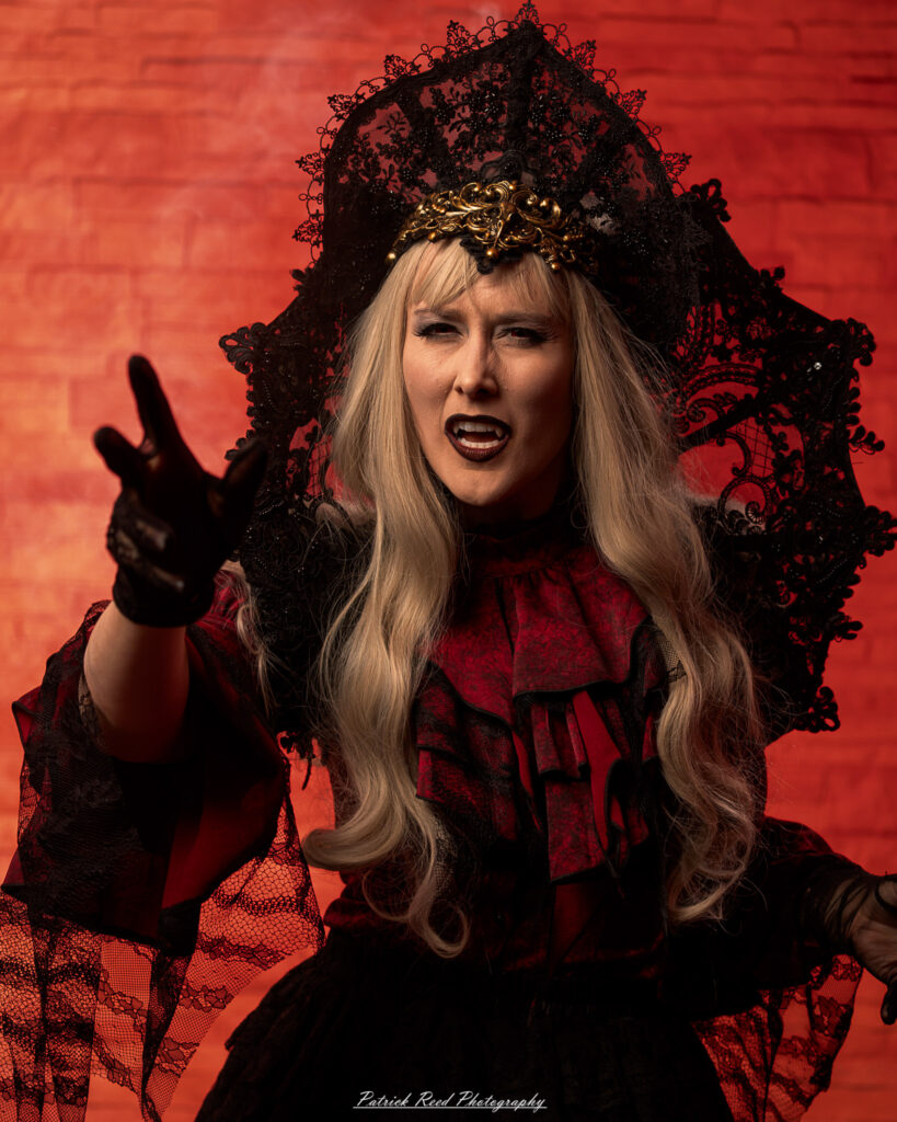 Alt text: Portrait of a woman in vampire cosplay named Lady Thorn, photographed on June 28, 2025. She is posed against a vivid red background, styled with gothic elements and dramatic expression.