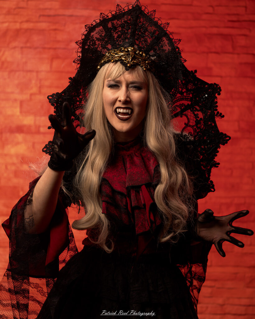 Alt text: Portrait of a woman in vampire cosplay named Lady Thorn, photographed on June 28, 2025. She is posed against a vivid red background, styled with gothic elements and dramatic expression.