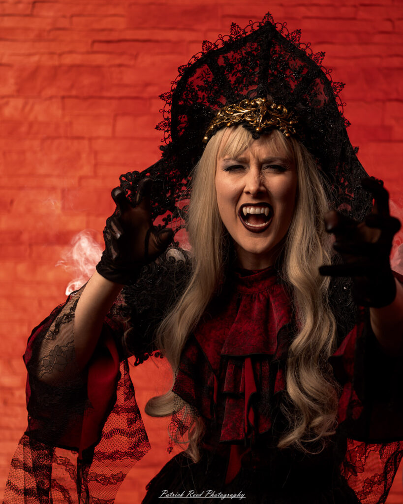 Alt text: Portrait of a woman in vampire cosplay named Lady Thorn, photographed on June 28, 2025. She is posed against a vivid red background, styled with gothic elements and dramatic expression.