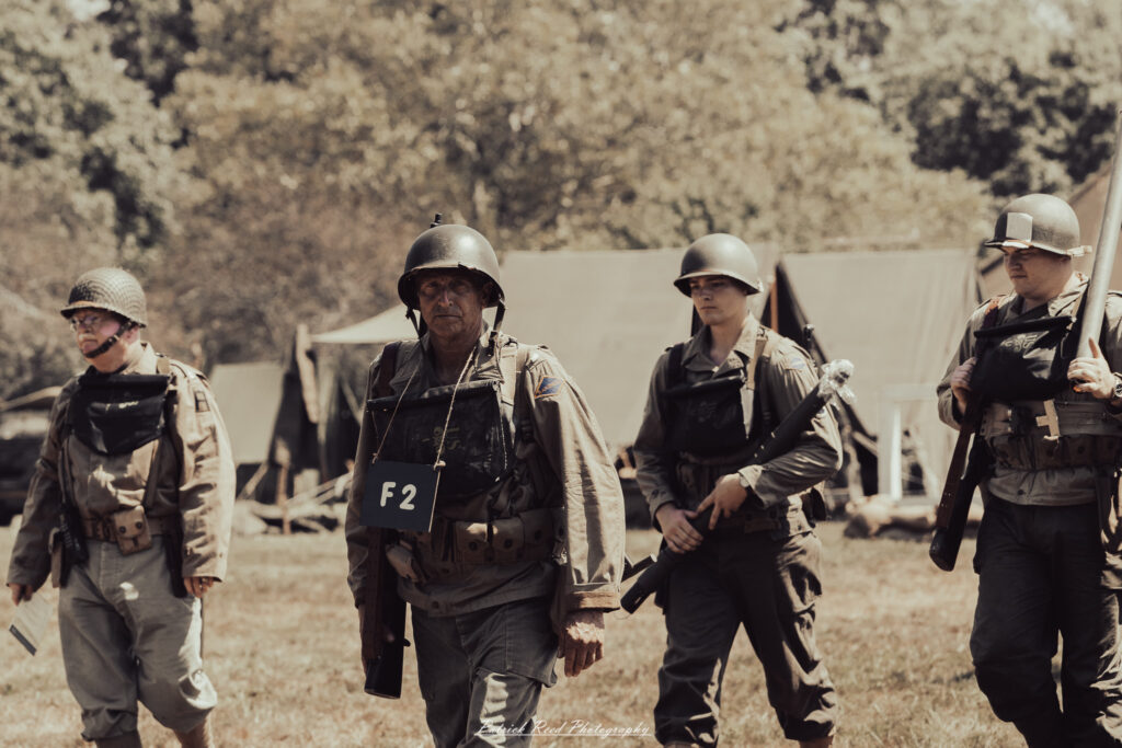A group of World War II reenactors in period-accurate uniforms—American, German, and French Resistance—on a sandy shore during the D-Day Conneaut 2025 reenactment at Conneaut Township Park, Ohio.