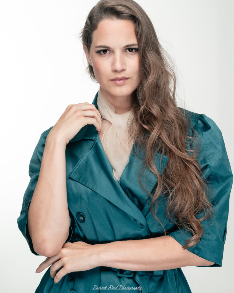 A dramatic studio portrait of woman wearing a stylish teal trench coat, photographed on August 23, 2025, with moody, directional lighting for a bold and modern look.