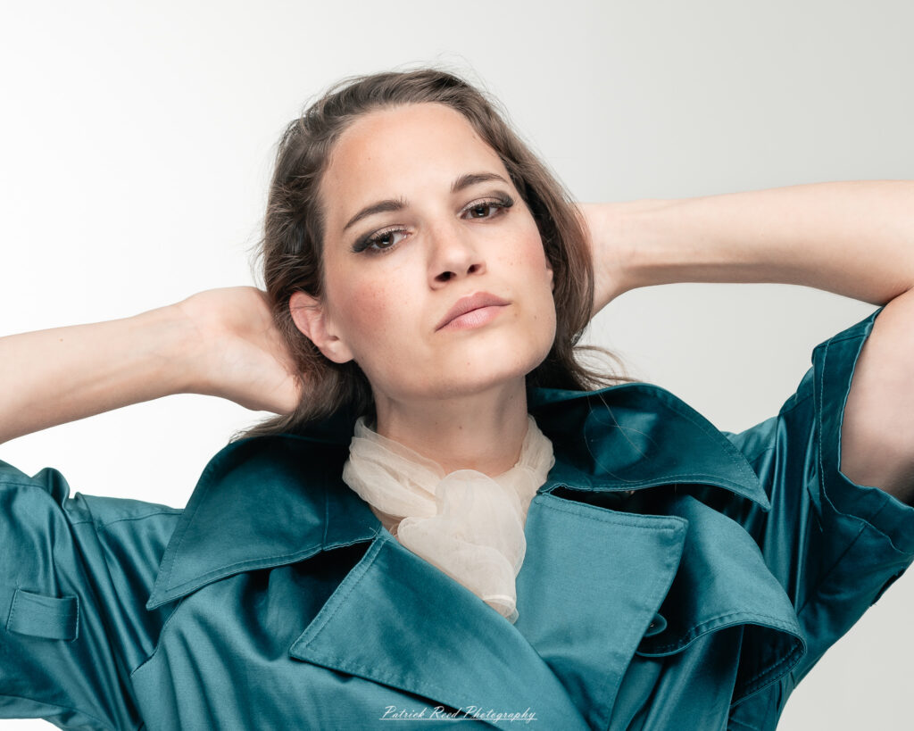 A dramatic studio portrait of woman wearing a stylish teal trench coat, photographed on August 23, 2025, with moody, directional lighting for a bold and modern look.