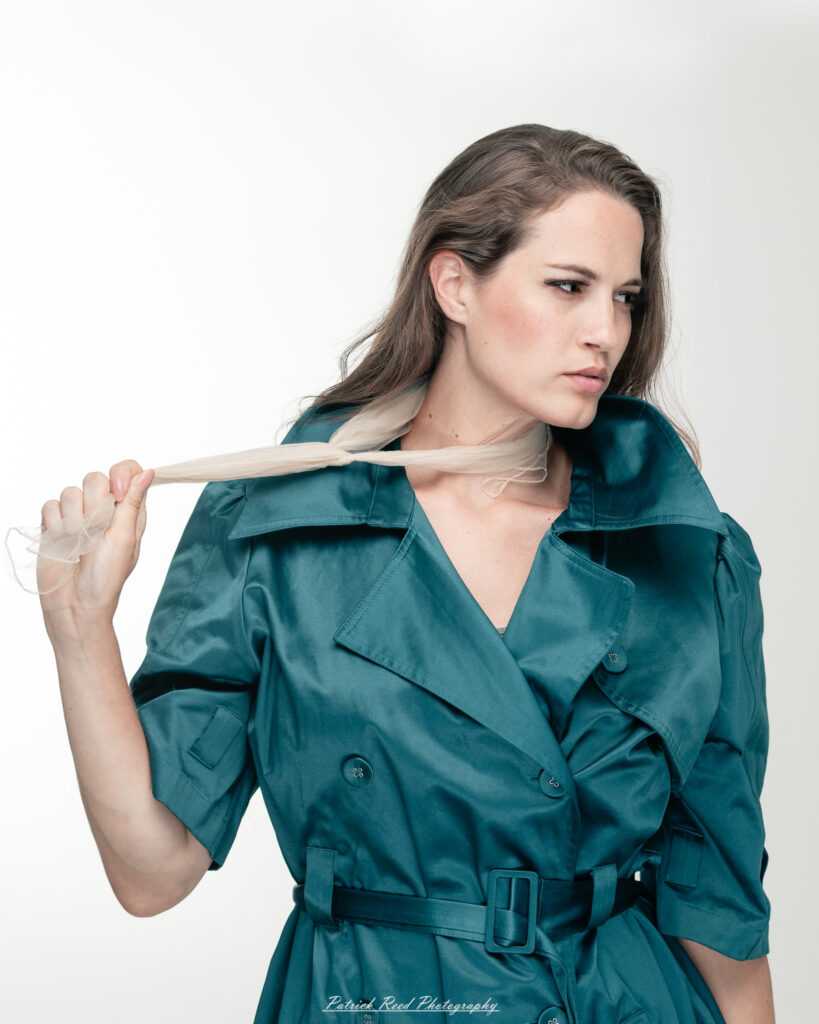 A dramatic studio portrait of woman wearing a stylish teal trench coat, photographed on August 23, 2025, with moody, directional lighting for a bold and modern look.