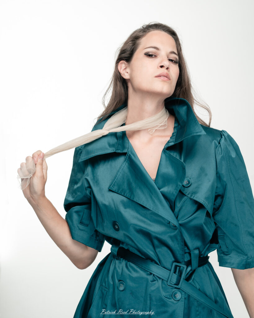 A dramatic studio portrait of woman wearing a stylish teal trench coat, photographed on August 23, 2025, with moody, directional lighting for a bold and modern look.