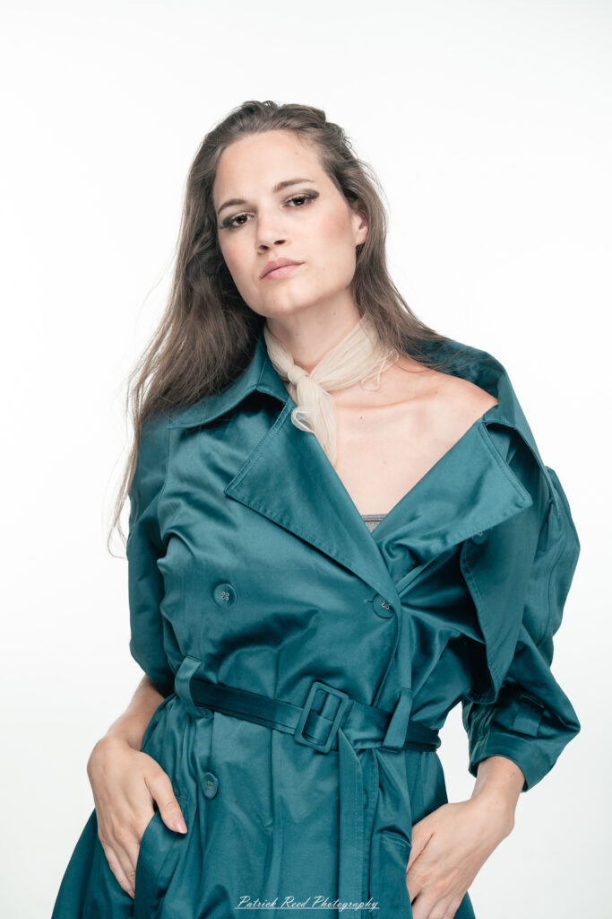 A dramatic studio portrait of woman wearing a stylish teal trench coat, photographed on August 23, 2025, with moody, directional lighting for a bold and modern look.