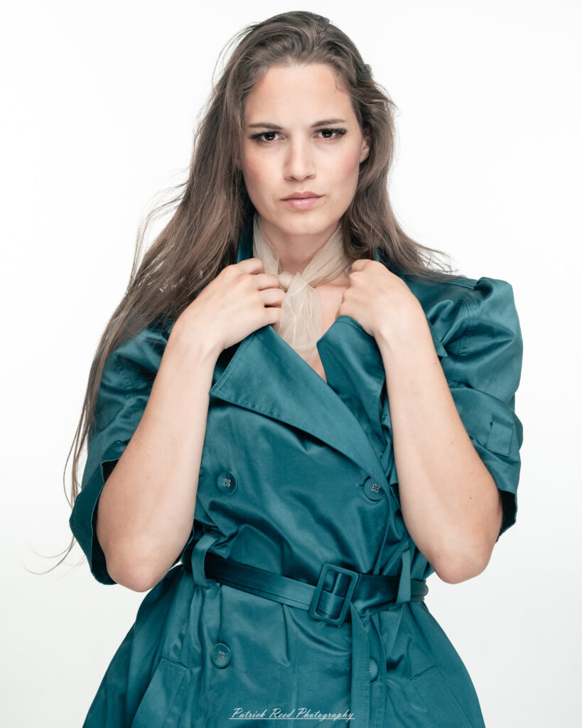 A dramatic studio portrait of woman wearing a stylish teal trench coat, photographed on August 23, 2025, with moody, directional lighting for a bold and modern look.
