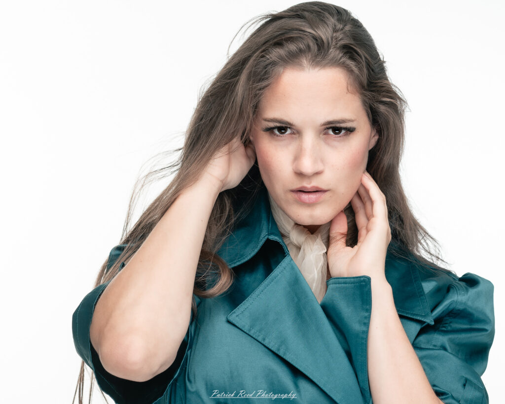 A dramatic studio portrait of woman wearing a stylish teal trench coat, photographed on August 23, 2025, with moody, directional lighting for a bold and modern look.