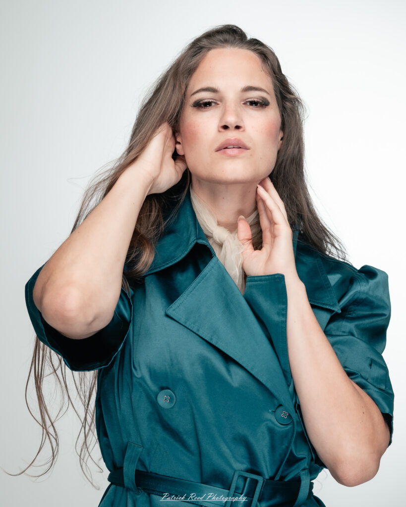 A dramatic studio portrait of woman wearing a stylish teal trench coat, photographed on August 23, 2025, with moody, directional lighting for a bold and modern look.