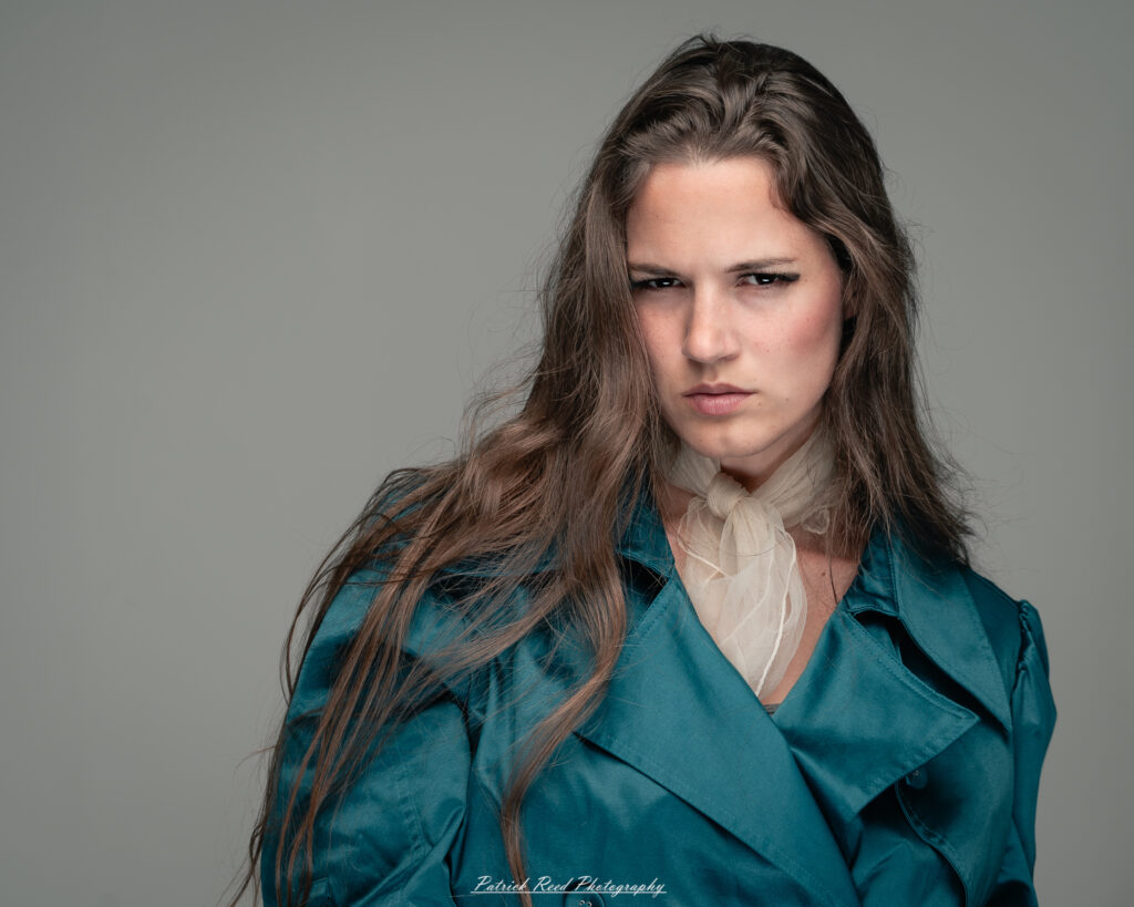 A dramatic studio portrait of woman wearing a stylish teal trench coat, photographed on August 23, 2025, with moody, directional lighting for a bold and modern look.