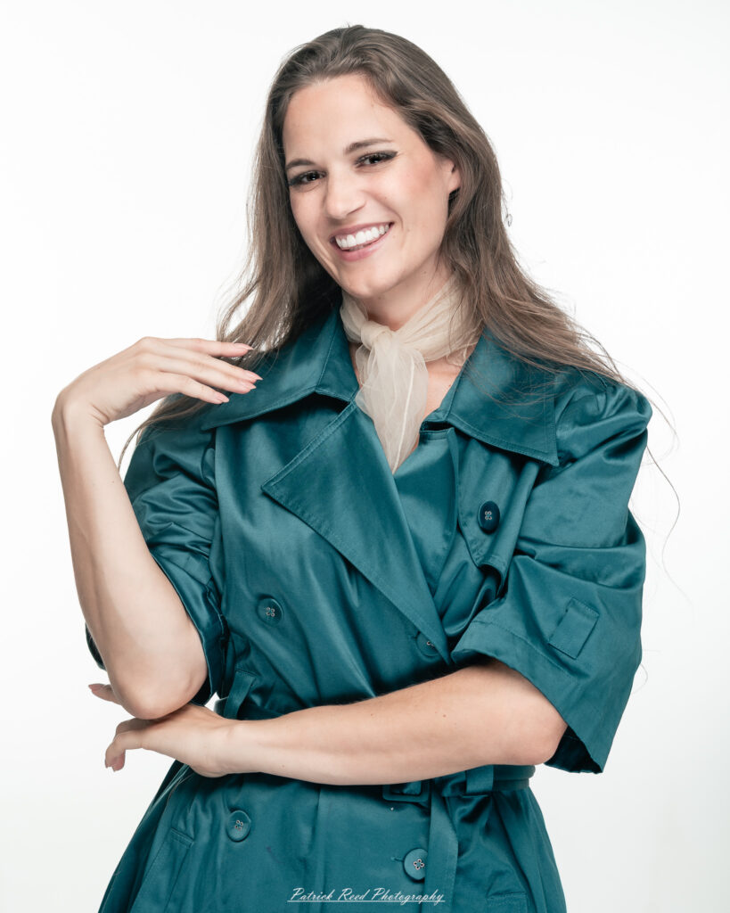A dramatic studio portrait of woman wearing a stylish teal trench coat, photographed on August 23, 2025, with moody, directional lighting for a bold and modern look.