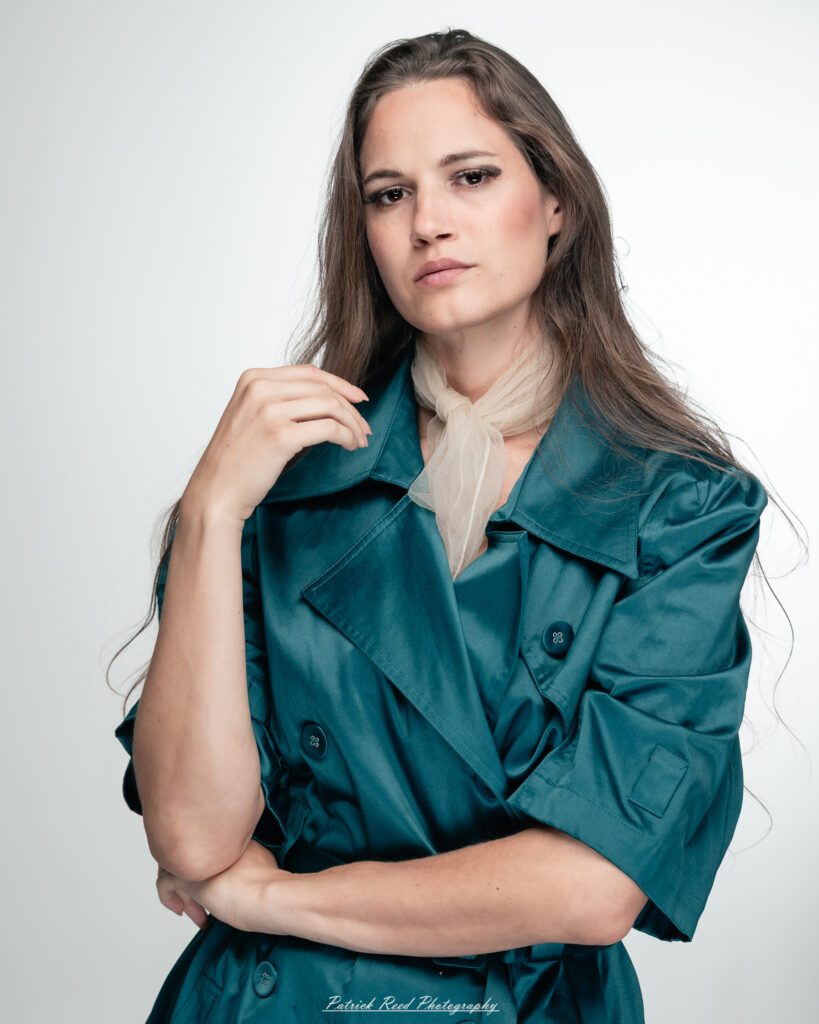 A dramatic studio portrait of woman wearing a stylish teal trench coat, photographed on August 23, 2025, with moody, directional lighting for a bold and modern look.