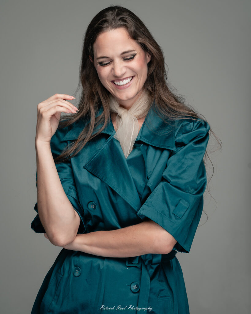 A dramatic studio portrait of woman wearing a stylish teal trench coat, photographed on August 23, 2025, with moody, directional lighting for a bold and modern look.