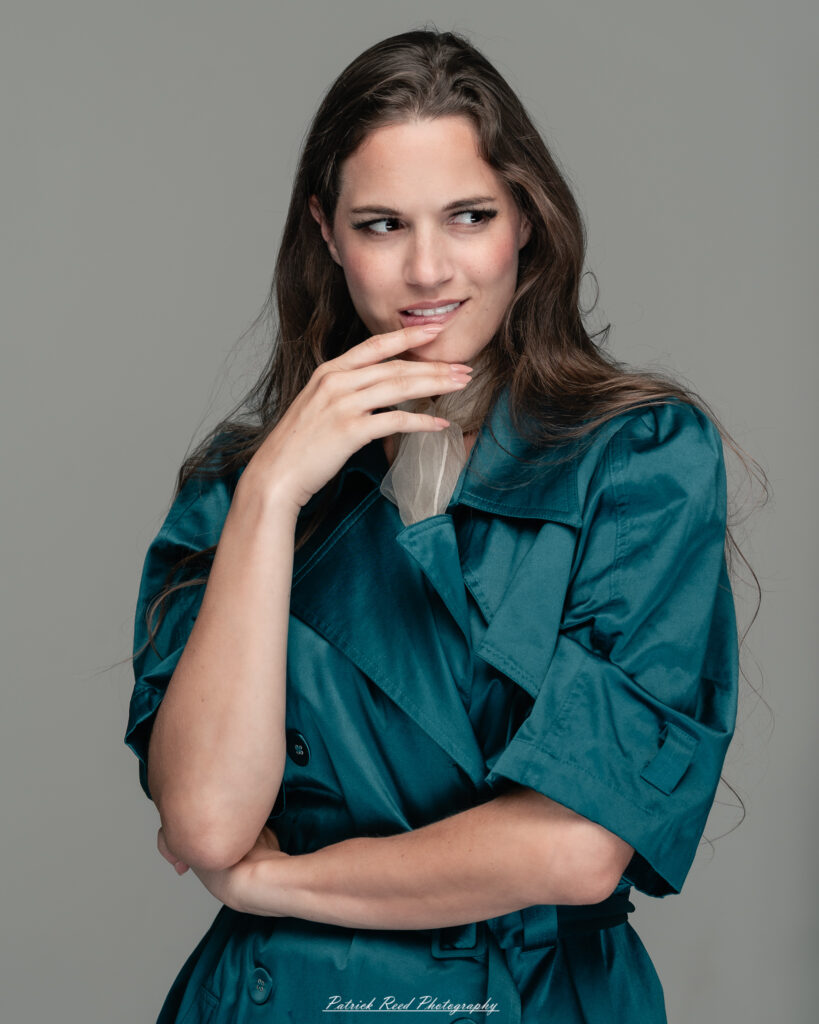 A dramatic studio portrait of woman wearing a stylish teal trench coat, photographed on August 23, 2025, with moody, directional lighting for a bold and modern look.