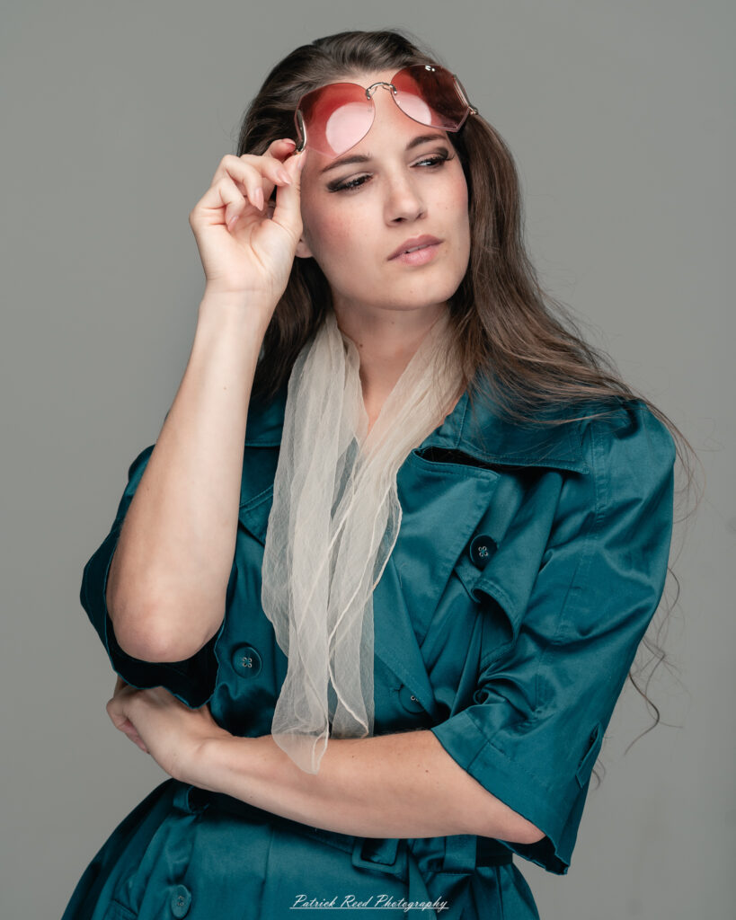 A dramatic studio portrait of woman wearing a stylish teal trench coat, photographed on August 23, 2025, with moody, directional lighting for a bold and modern look.