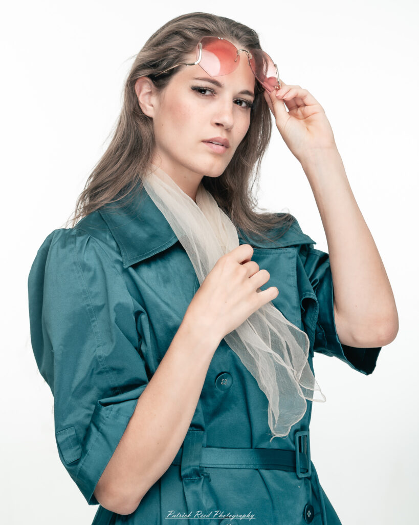 A dramatic studio portrait of woman wearing a stylish teal trench coat, photographed on August 23, 2025, with moody, directional lighting for a bold and modern look.