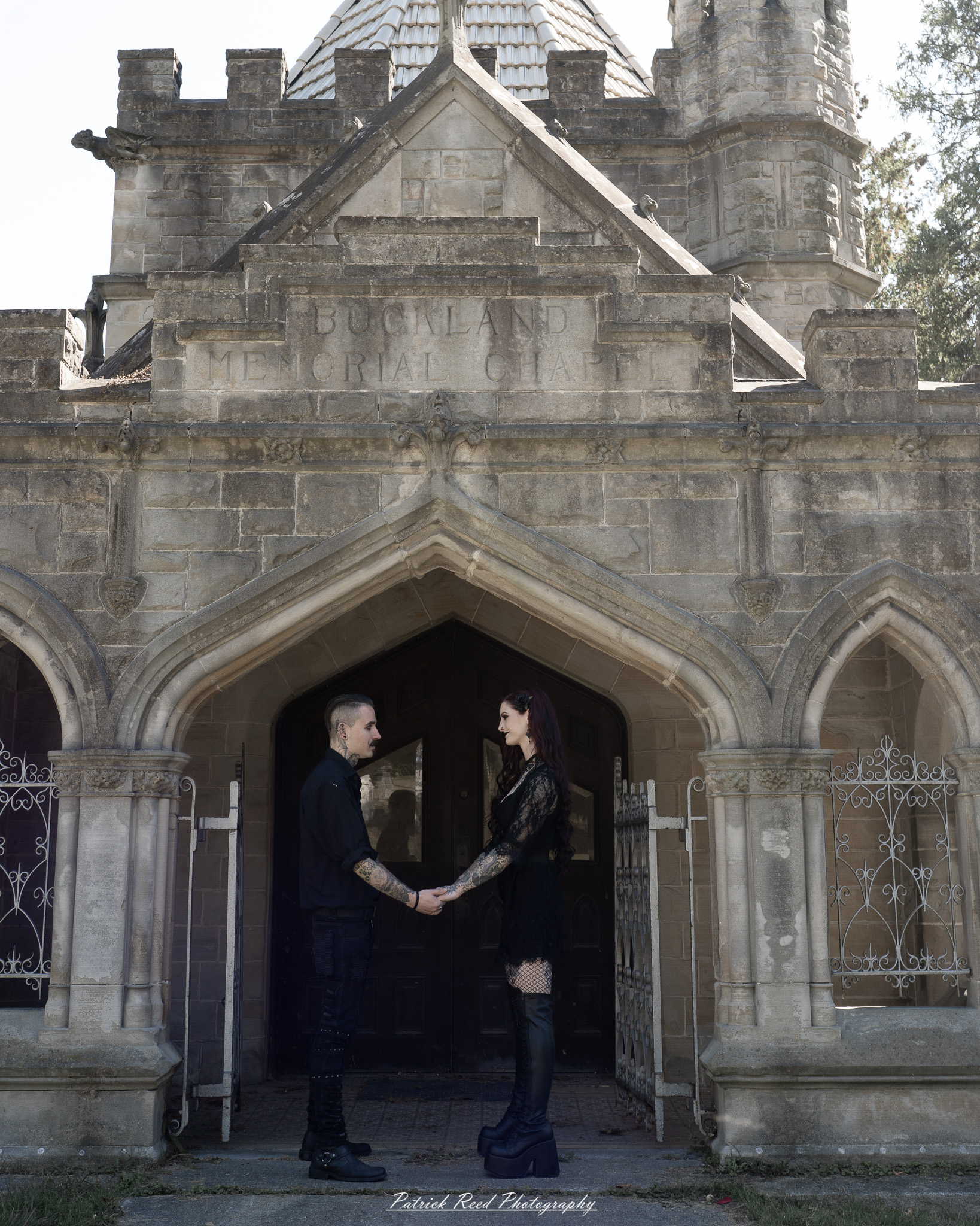 A gothic-themed portrait of a model named Minerva with a partner, photographed in a cemetery on September 28, 2025, featuring dark attire, moody lighting, and dramatic composition.