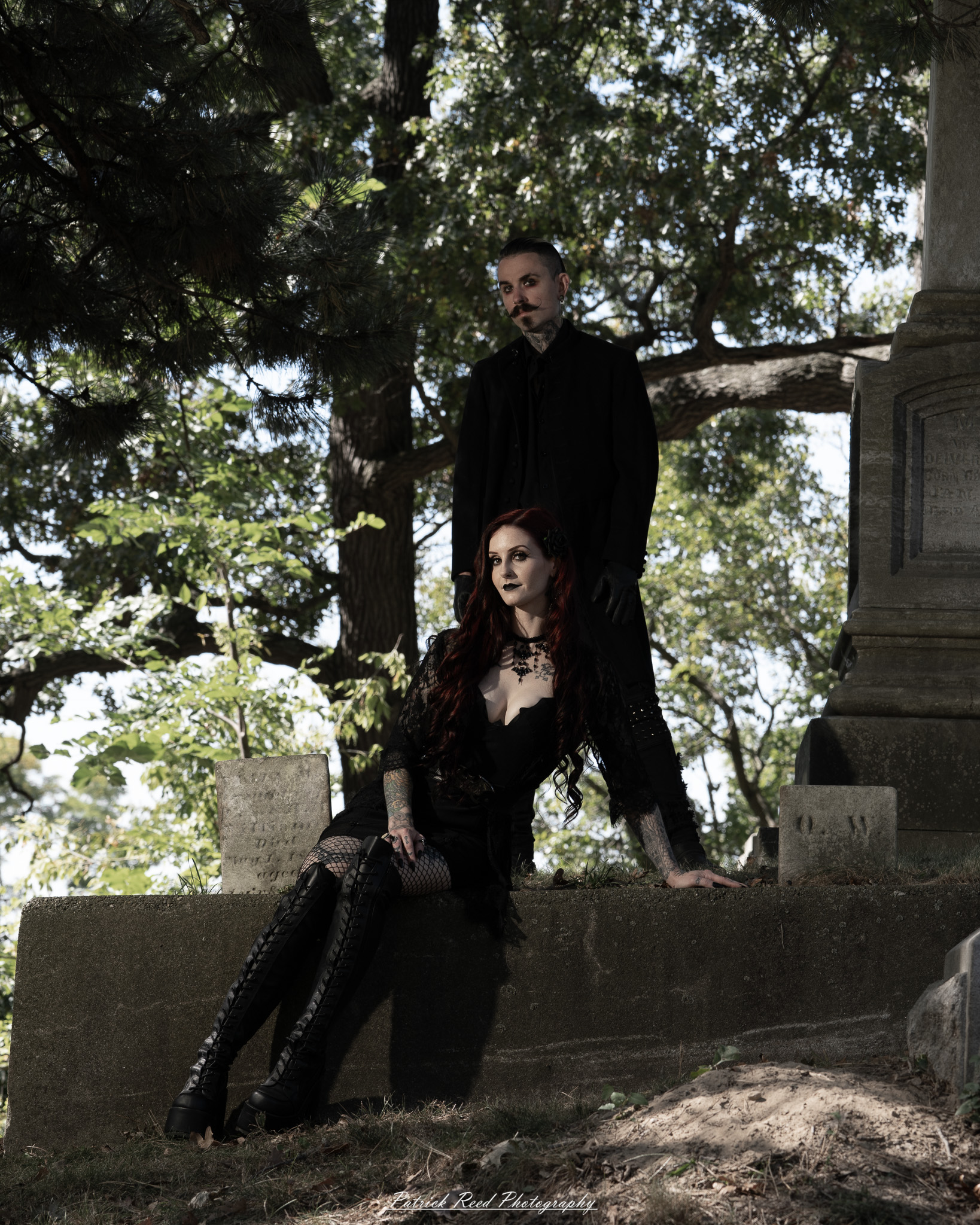 A gothic-themed portrait of a model named Minerva with a partner, photographed in a cemetery on September 28, 2025, featuring dark attire, moody lighting, and dramatic composition.