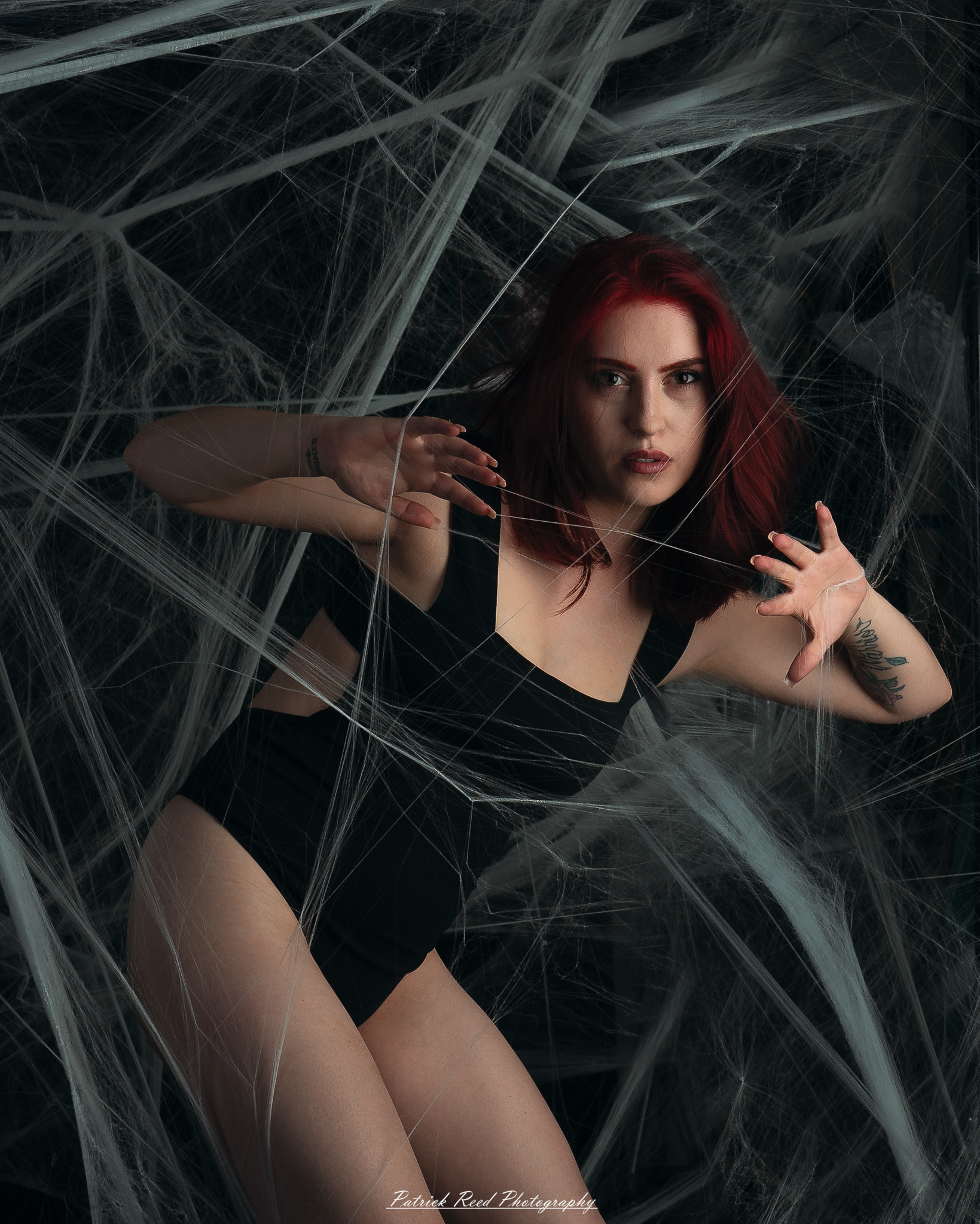 A portrait of a woman named Jinx surrounded by artificial spiderwebs, photographed on October 29, 2025. The lighting is dramatic and moody, emphasizing the texture of the webs and her expression.