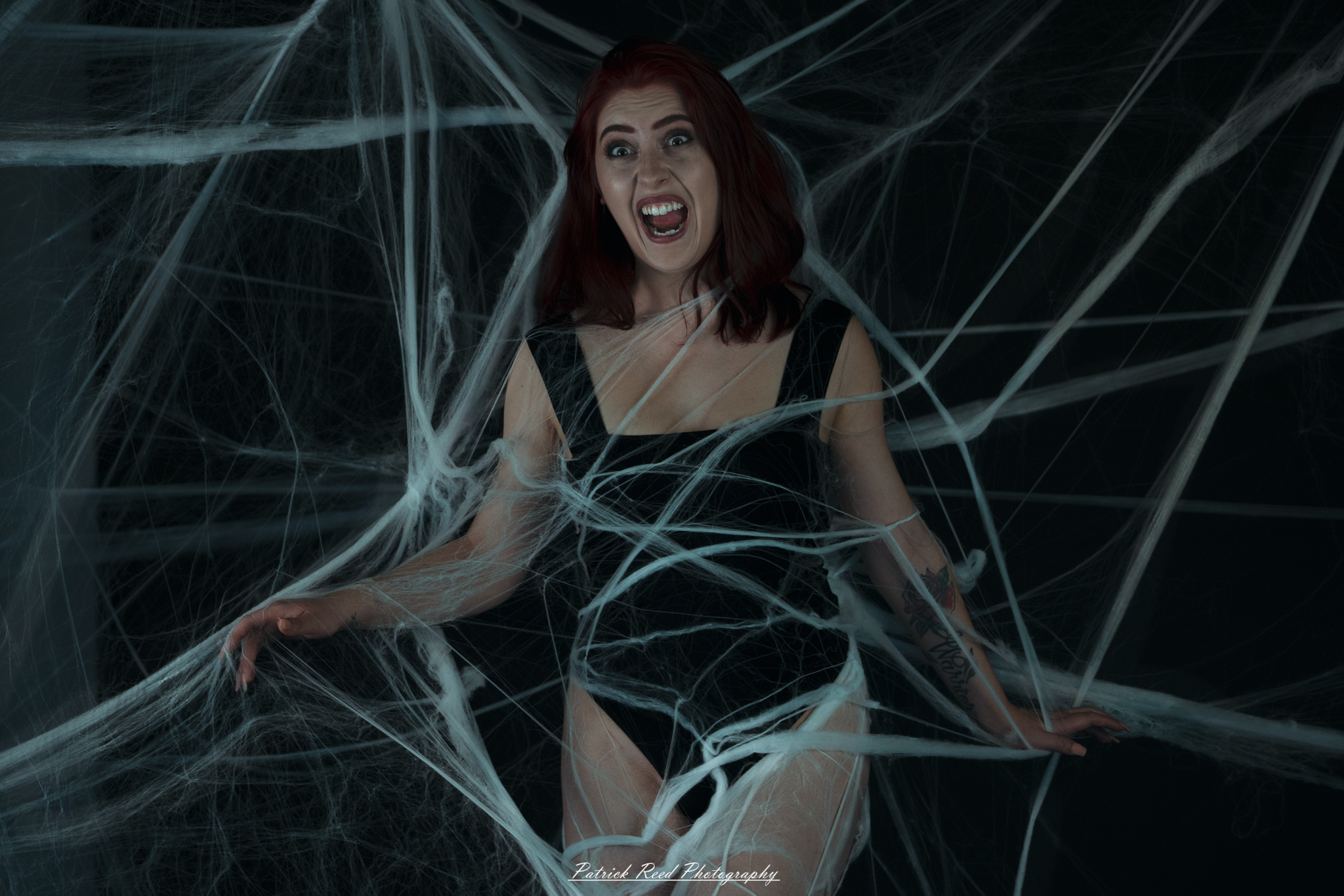 A portrait of a woman named Jinx surrounded by artificial spiderwebs, photographed on October 29, 2025. The lighting is dramatic and moody, emphasizing the texture of the webs and her expression.