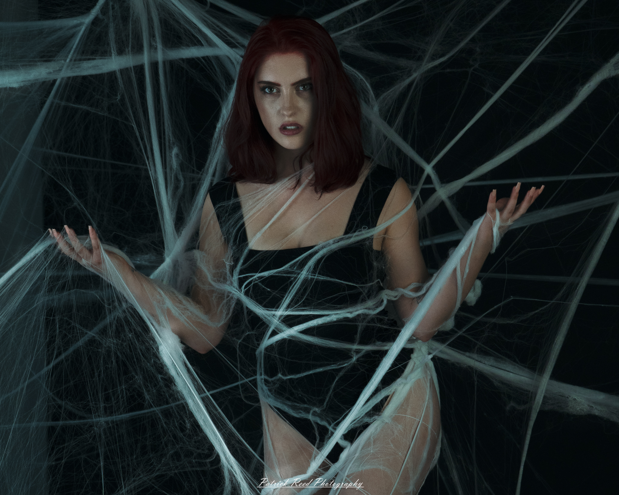 A portrait of a woman named Jinx surrounded by artificial spiderwebs, photographed on October 29, 2025. The lighting is dramatic and moody, emphasizing the texture of the webs and her expression.