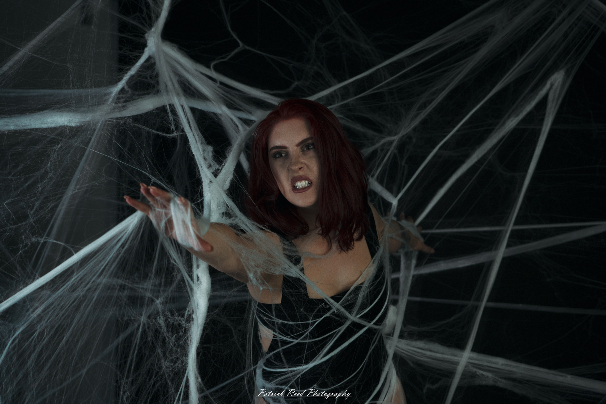 A portrait of a woman named Jinx surrounded by artificial spiderwebs, photographed on October 29, 2025. The lighting is dramatic and moody, emphasizing the texture of the webs and her expression.