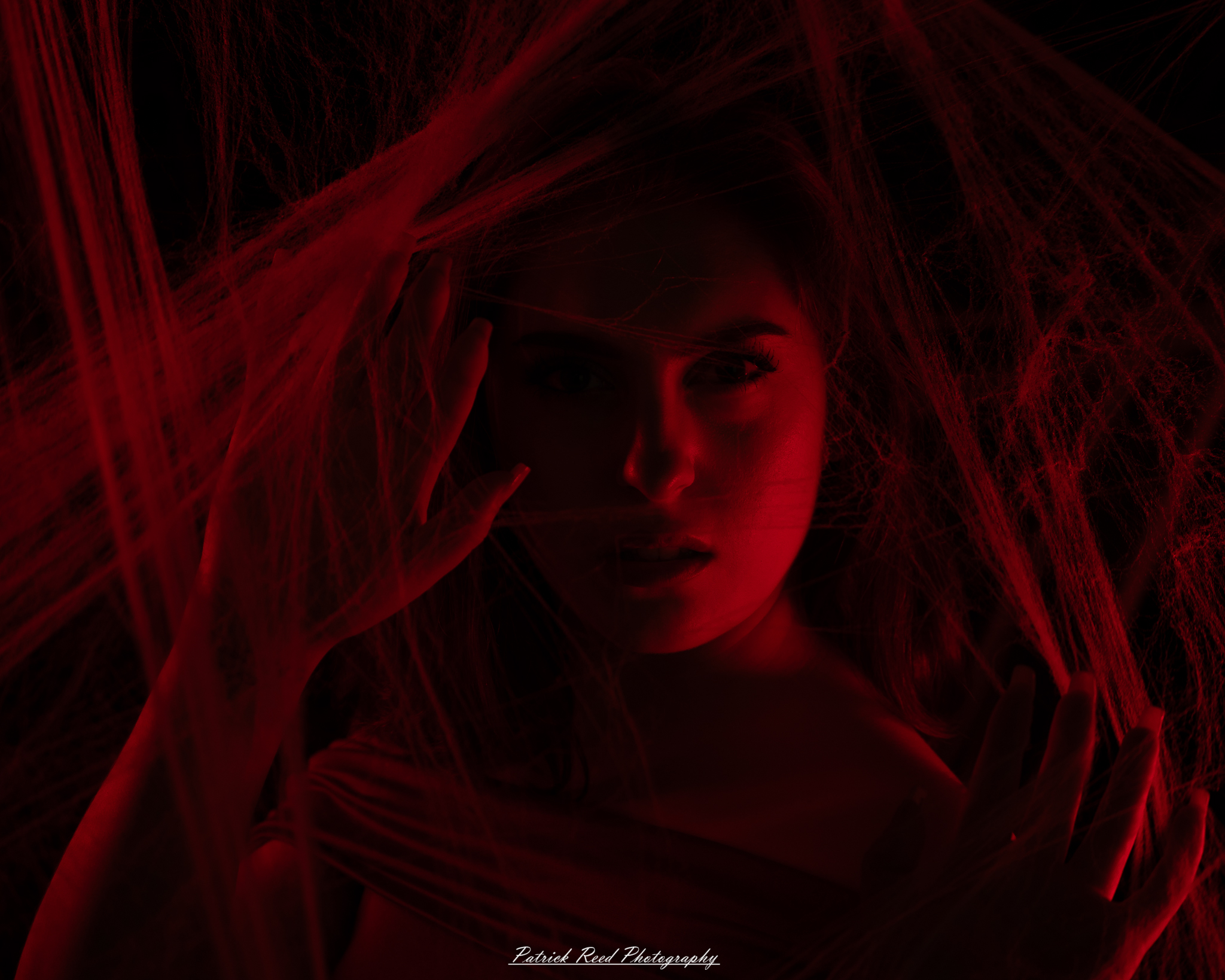 A portrait of a woman named Jinx surrounded by artificial spiderwebs, photographed on October 29, 2025. The lighting is dramatic and moody, emphasizing the texture of the webs and her expression.