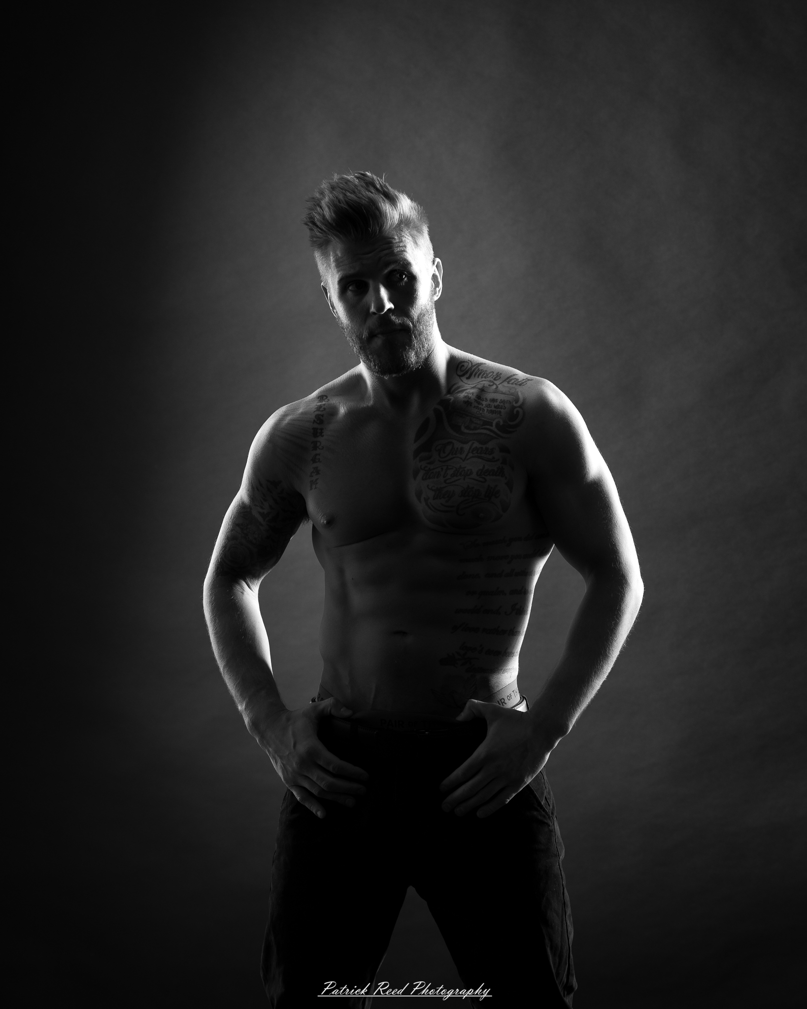 Black and white studio portrait of a shirtless male figure standing confidently with soft halo backlight