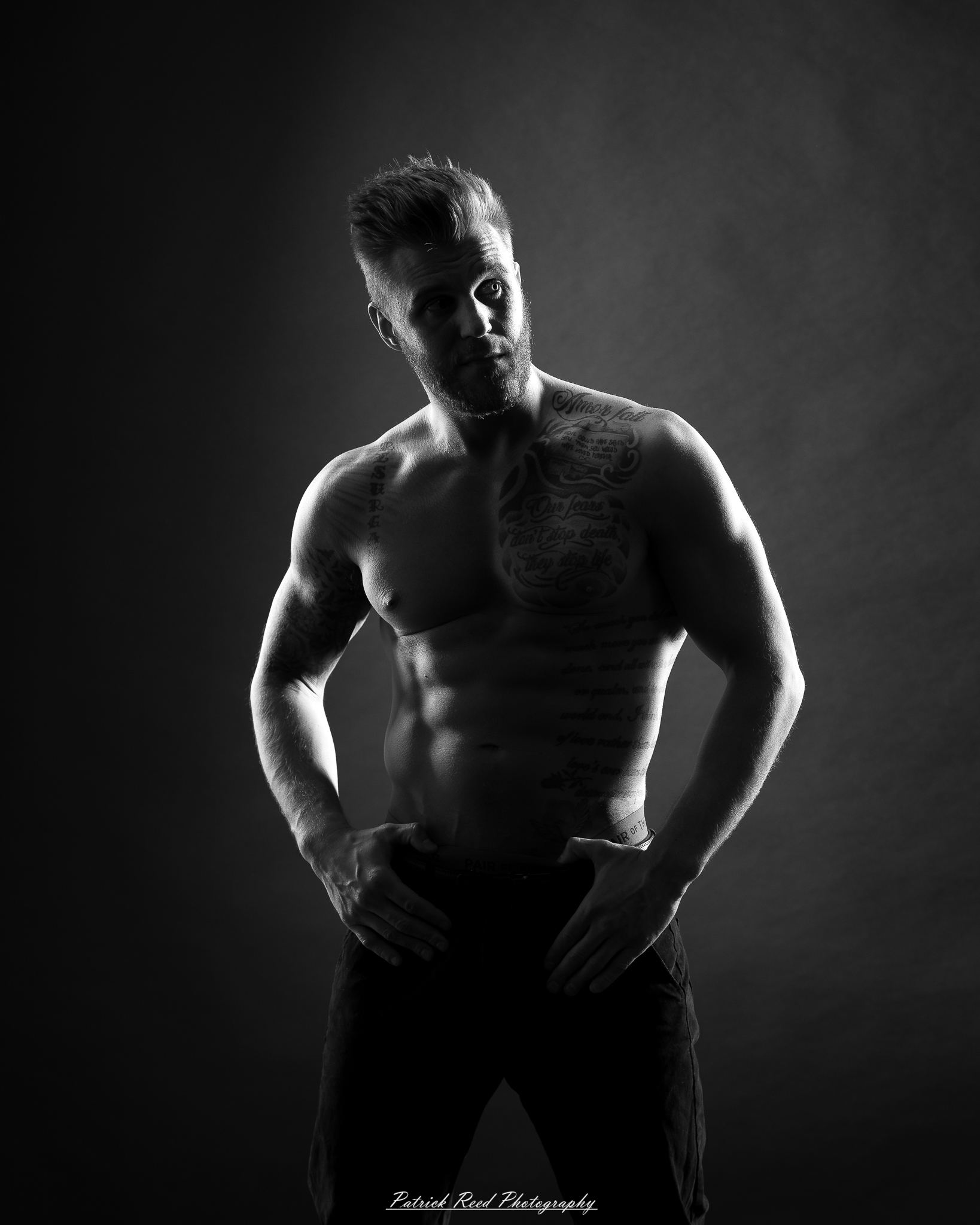 Black and white studio portrait of a shirtless male figure standing confidently with soft halo backlight
