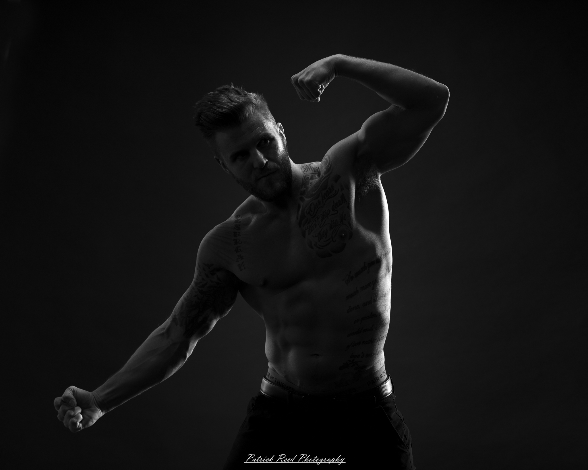 Black and white studio portrait of a shirtless male figure standing confidently with soft halo backlight