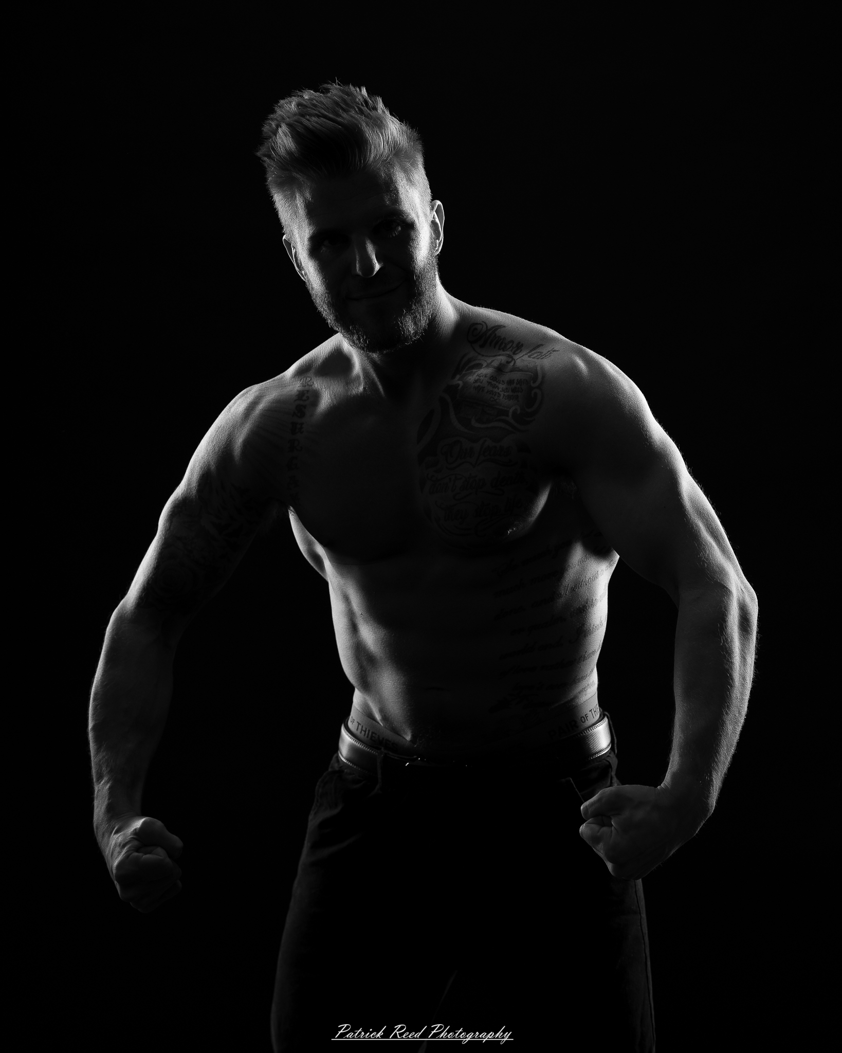 Black and white studio portrait of a shirtless male figure standing confidently with soft halo backlight