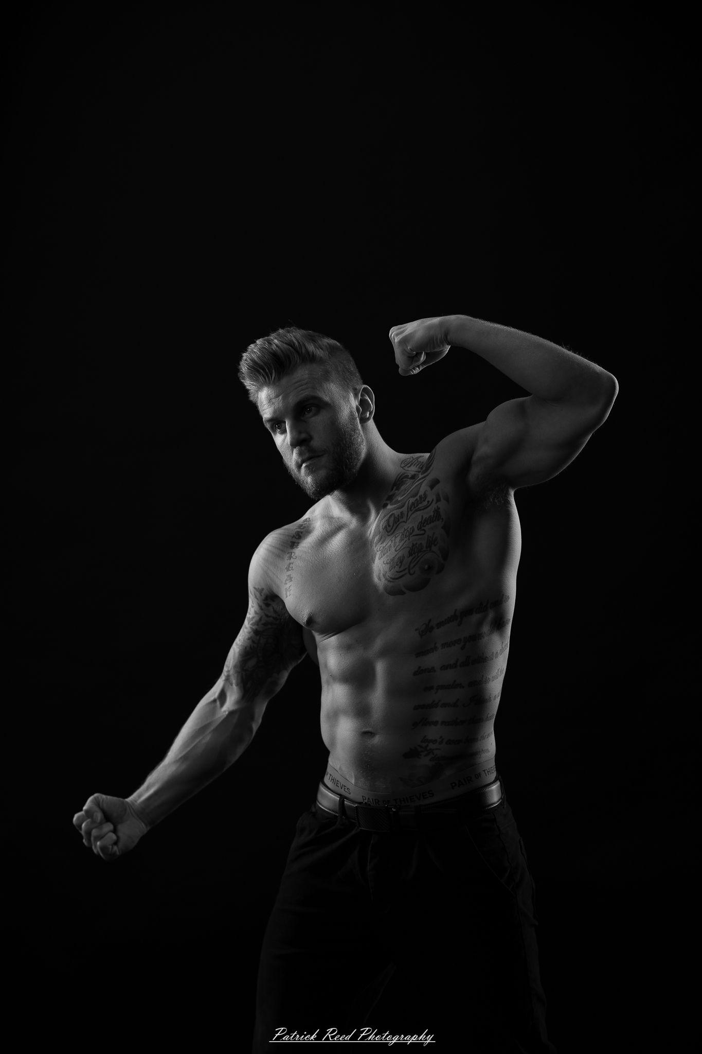 Black and white studio portrait of a shirtless male figure standing confidently with soft halo backlight