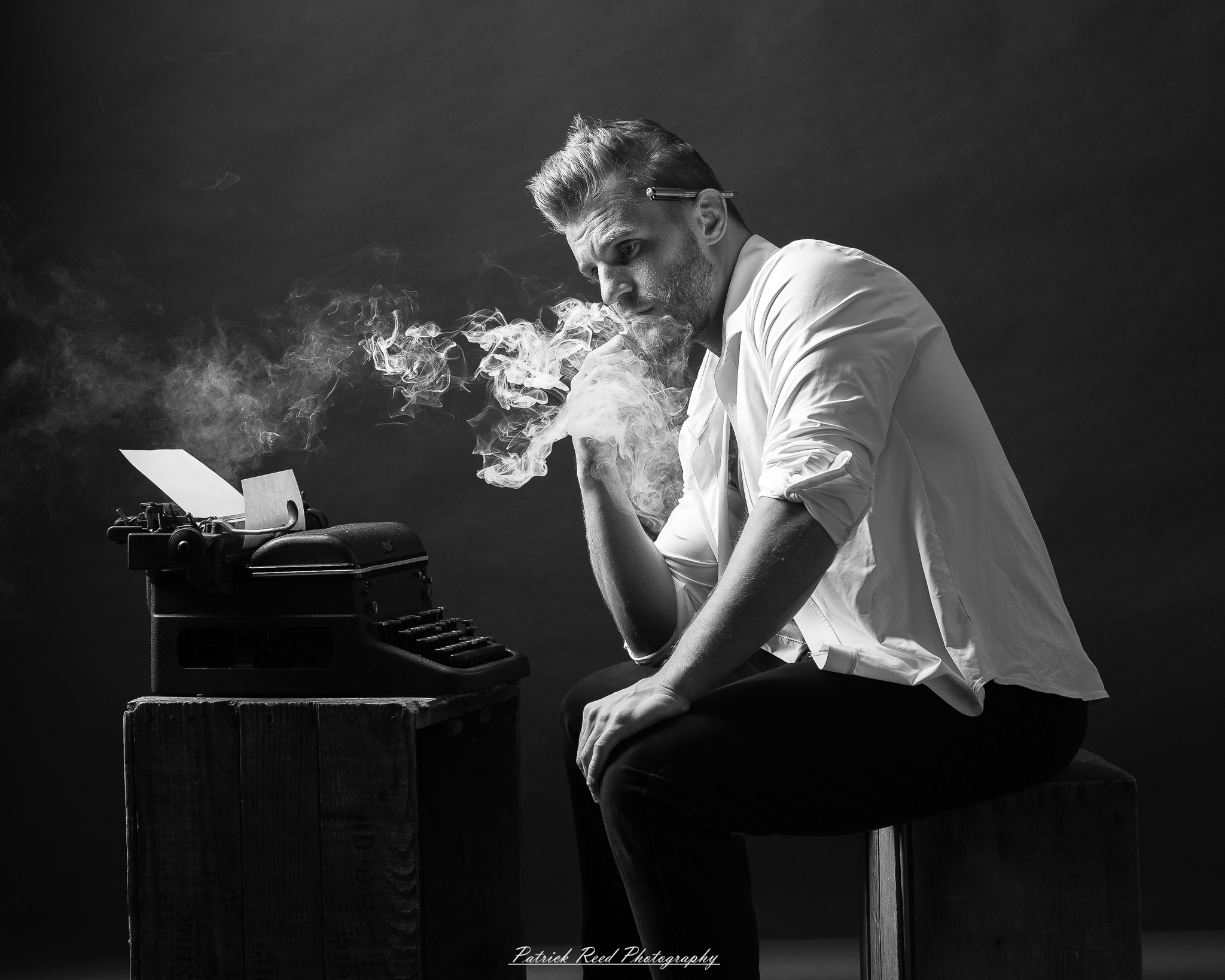 Black and white noir style portrait of a man sitting beside a vintage typewriter surrounded by drifting smoke