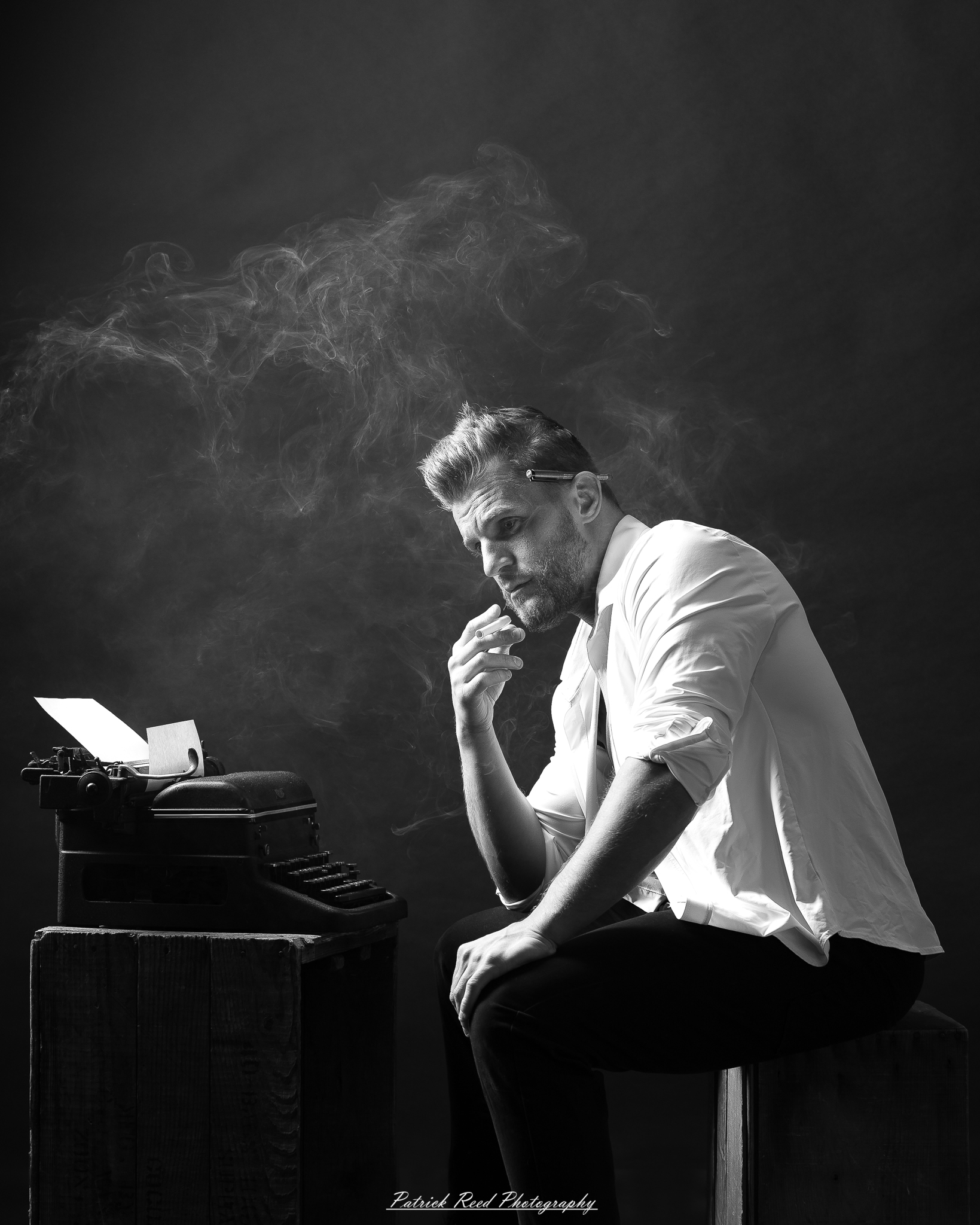 Black and white noir style portrait of a man sitting beside a vintage typewriter surrounded by drifting smoke