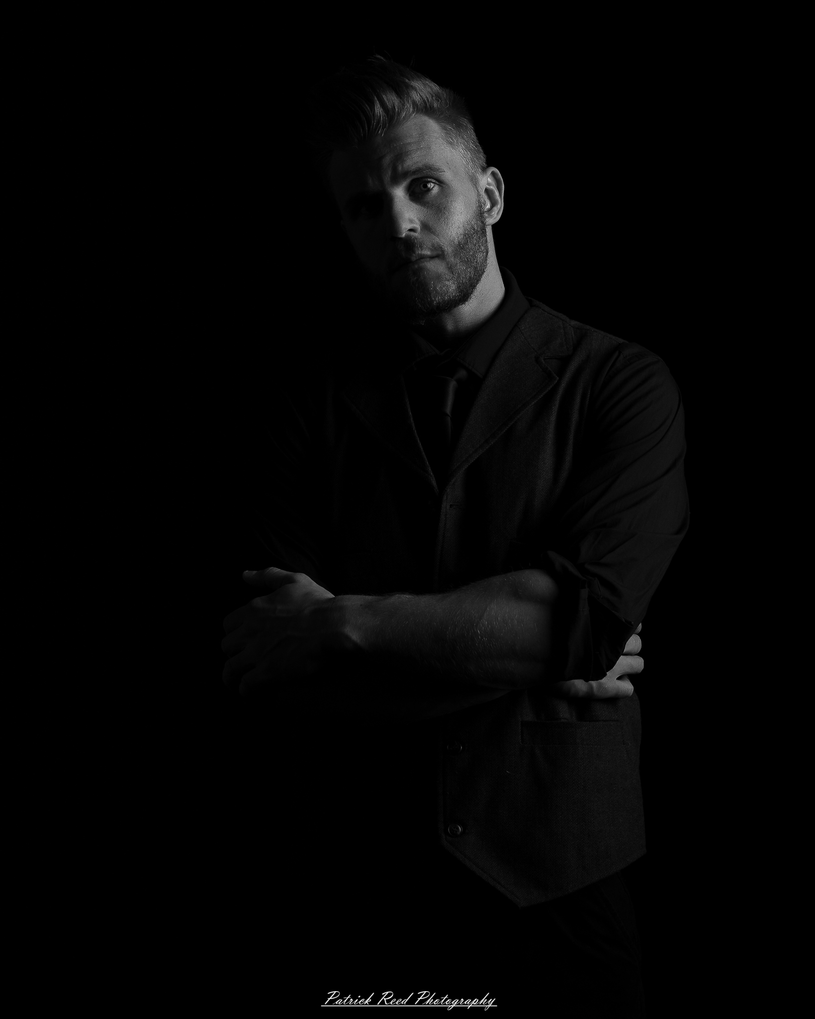 Black and white low-key portrait of a man standing with folded arms against a dark background with dramatic side lighting