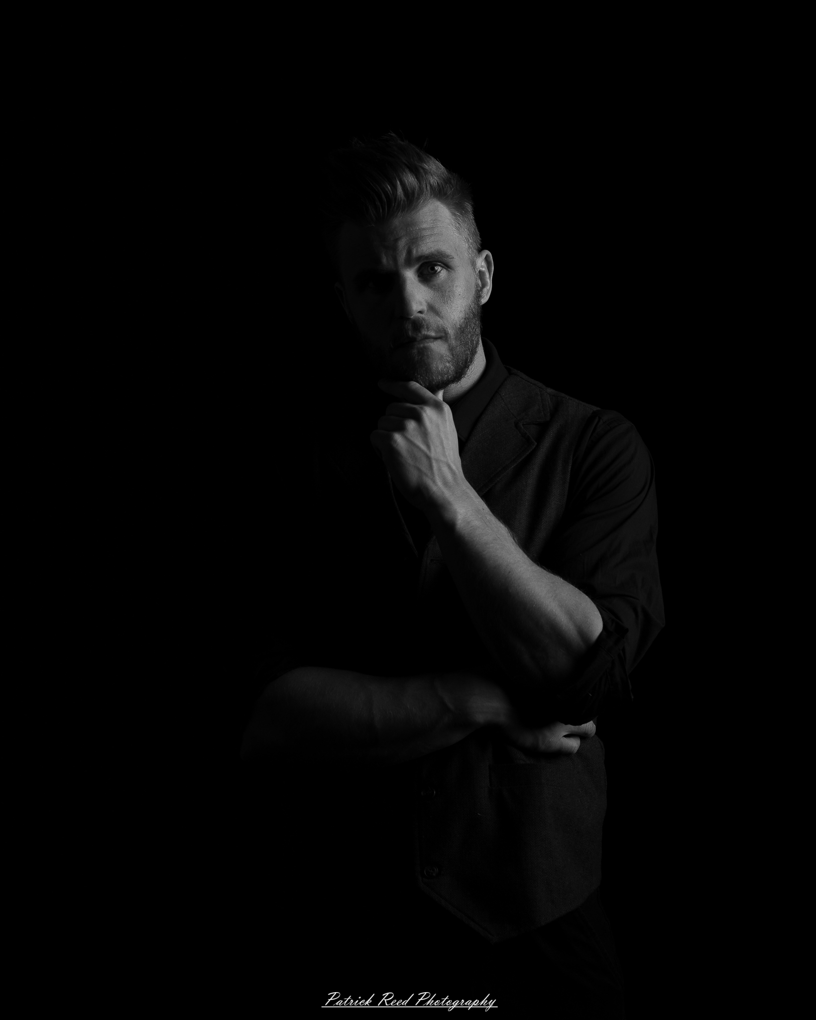 Black and white low-key portrait of a man standing with folded arms against a dark background with dramatic side lighting