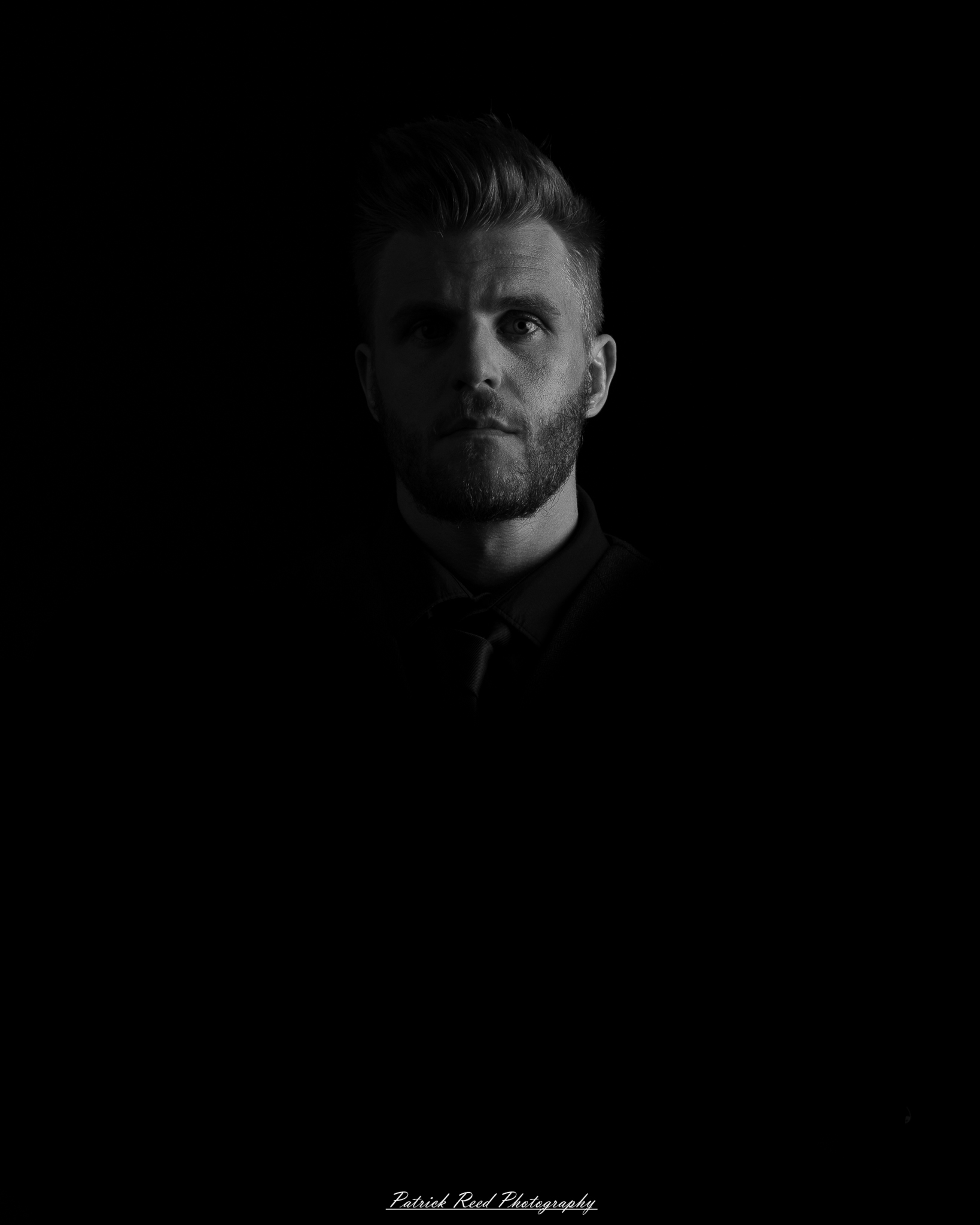 Black and white low-key portrait of a man standing with folded arms against a dark background with dramatic side lighting