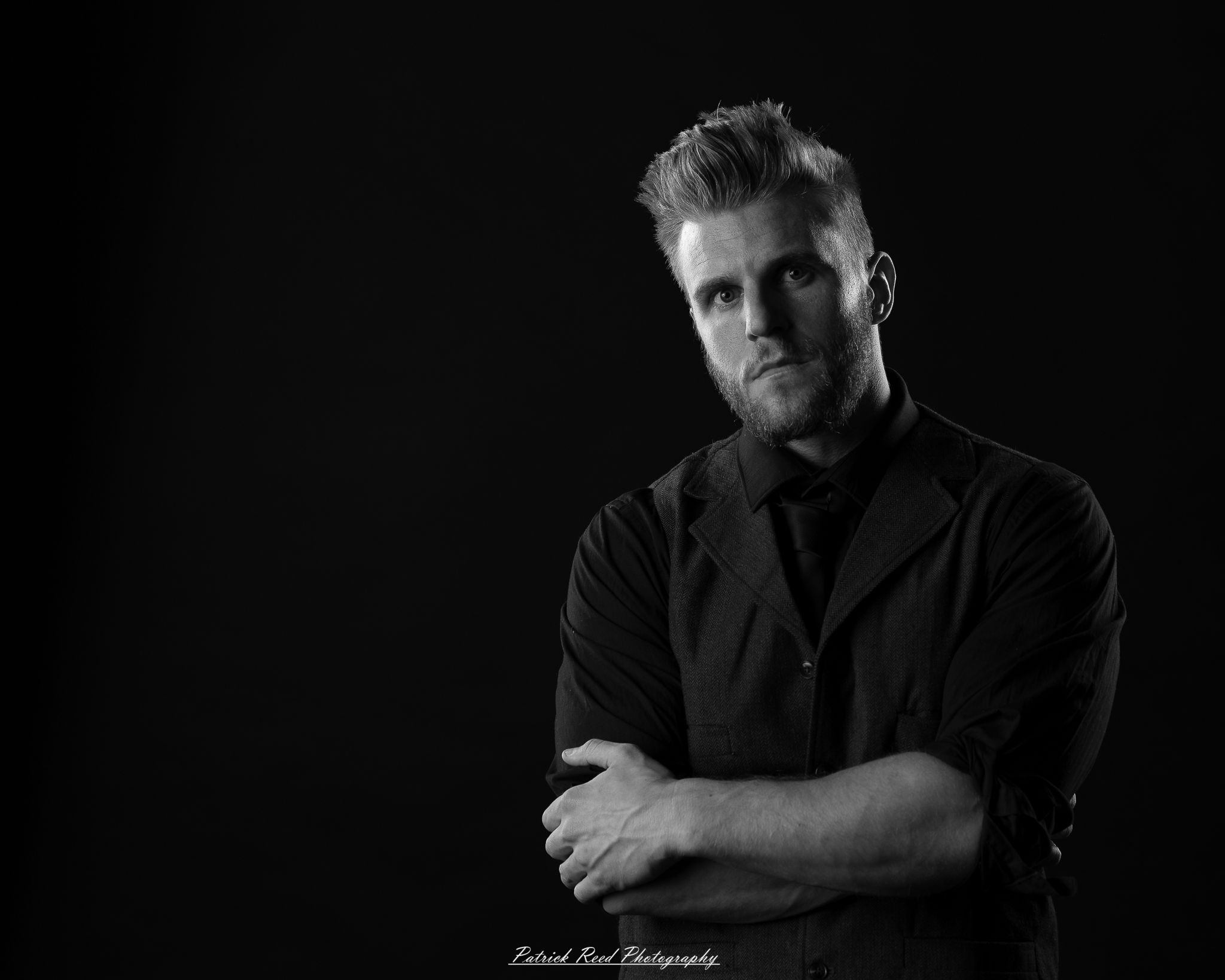 Black and white low-key portrait of a man standing with folded arms against a dark background with dramatic side lighting