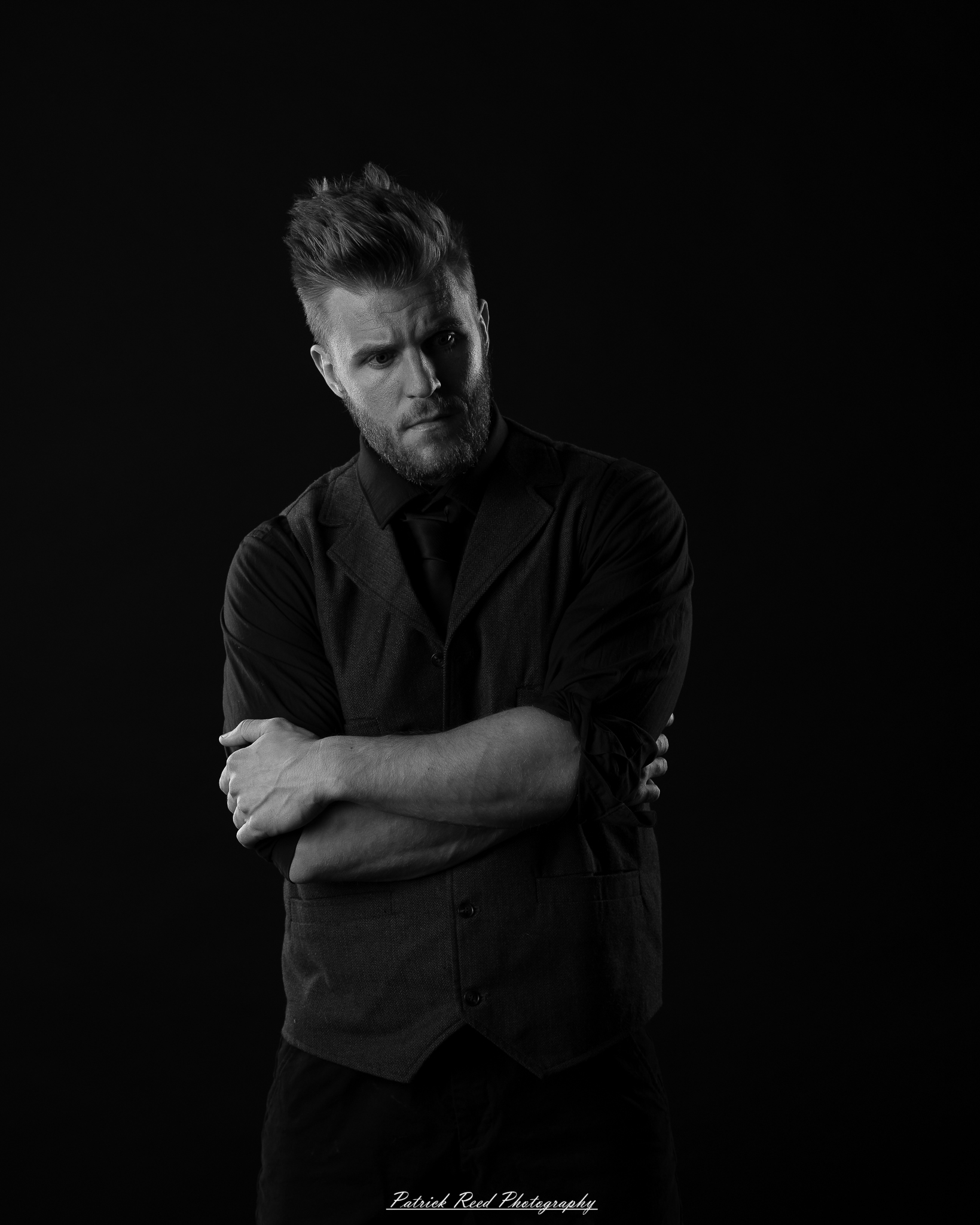 Black and white low-key portrait of a man standing with folded arms against a dark background with dramatic side lighting