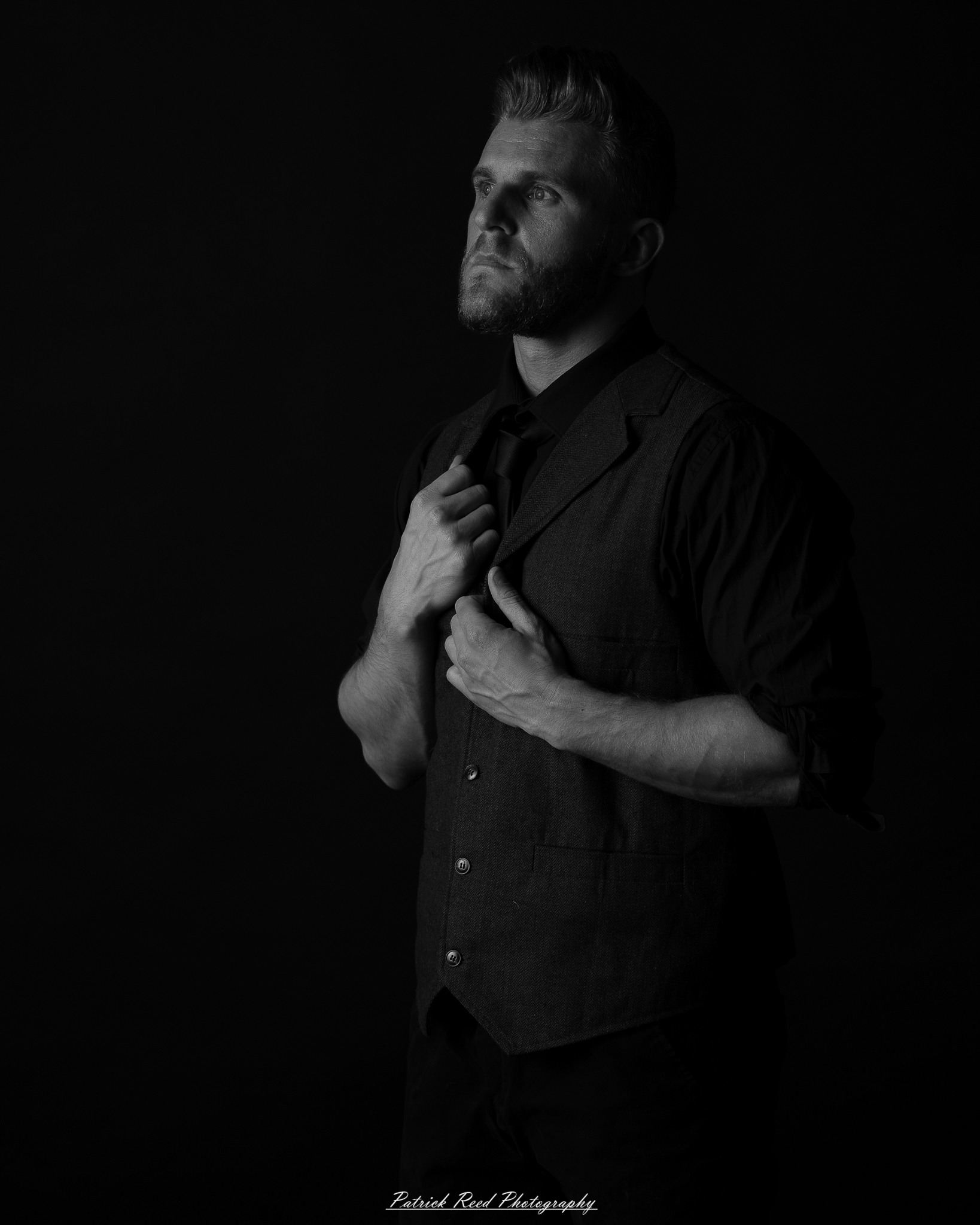 Black and white low-key portrait of a man standing with folded arms against a dark background with dramatic side lighting