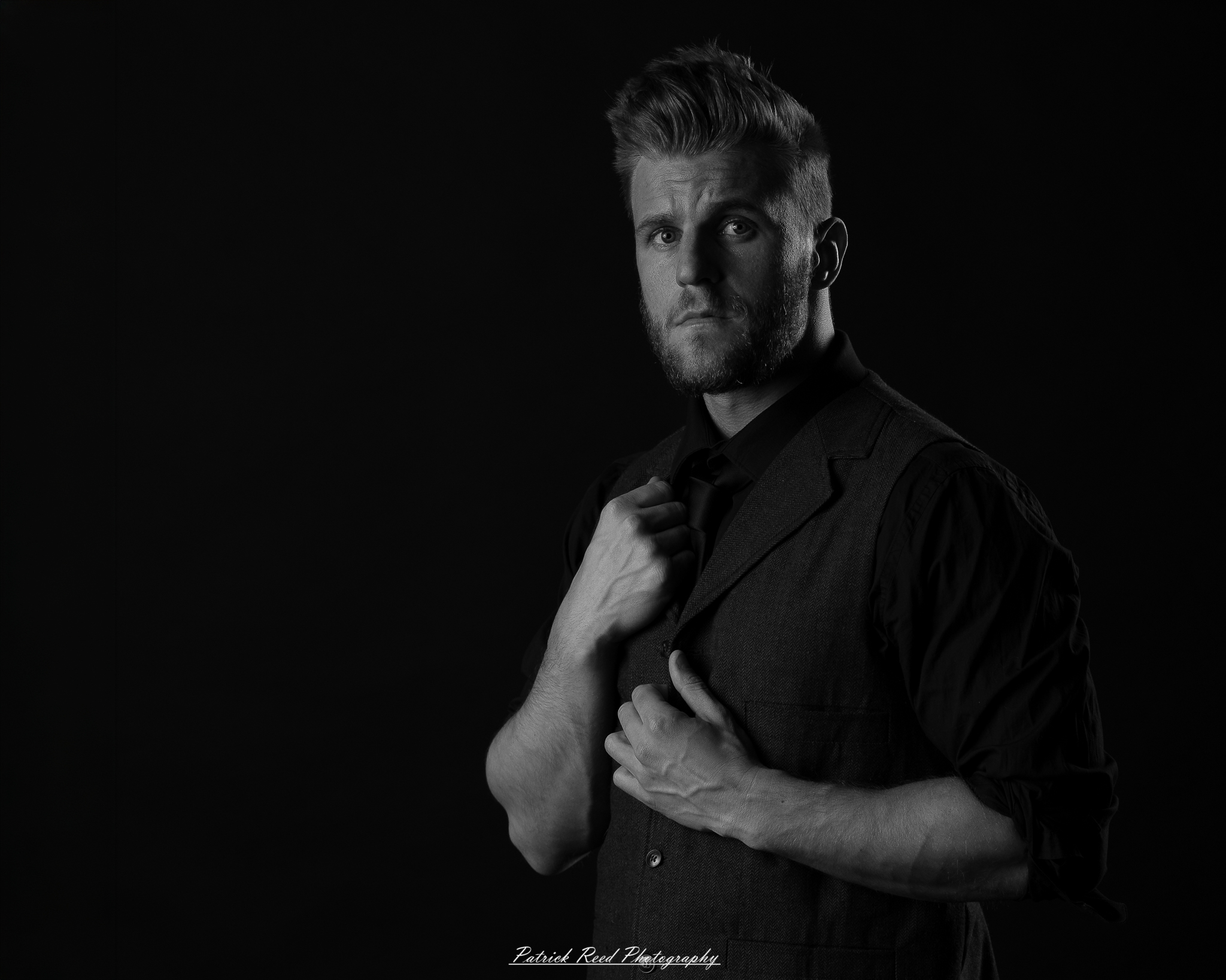 Black and white low-key portrait of a man standing with folded arms against a dark background with dramatic side lighting