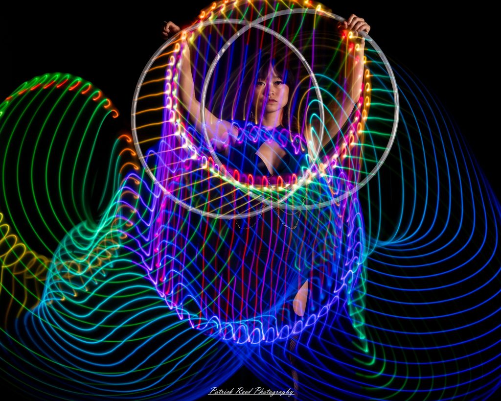 A studio portrait of a model surrounded by flowing light painting patterns created with long-exposure techniques, forming colorful arcs and motion trails against a black background.